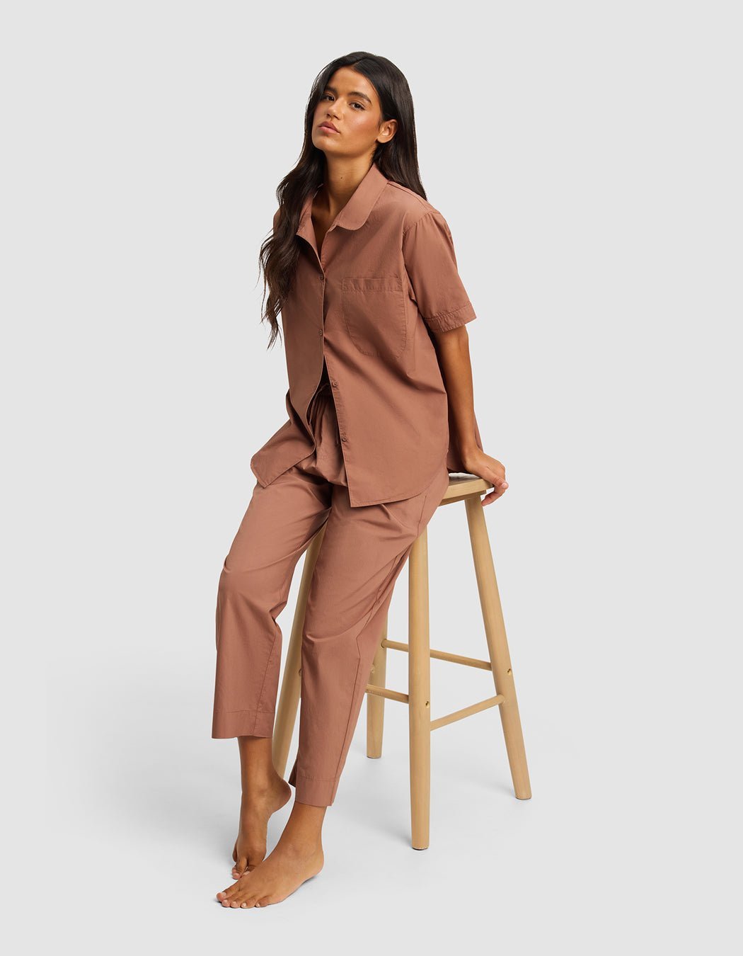 A woman with long dark hair sits barefoot on a wooden stool, wearing Cozy Earth's Women's Sutton Cropped Pajama Pant in brown with a matching short-sleeve button-up top, against a plain light gray background.