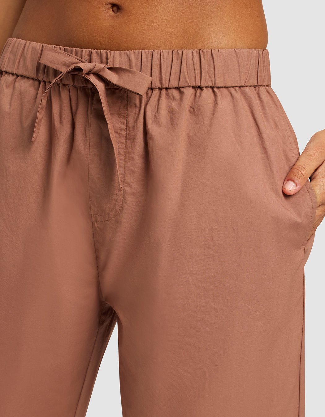 A close-up shows a person wearing Cozy Earth's Women's Sutton Cropped Pajama Pant in light brown, featuring a high waist, elastic waistband, drawstring tie, and their hand in one pocket.
