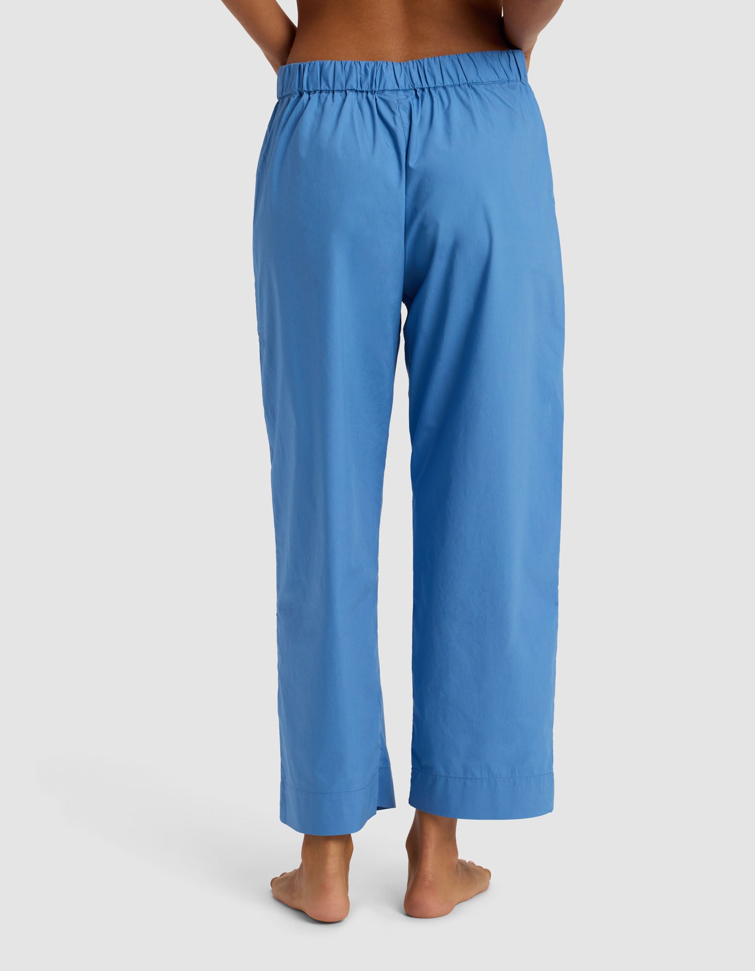 A person stands barefoot facing away, wearing Cozy Earth's Women's Sutton Cropped Pajama Pant in sky blue with an elastic waistband, set against a light gray background.