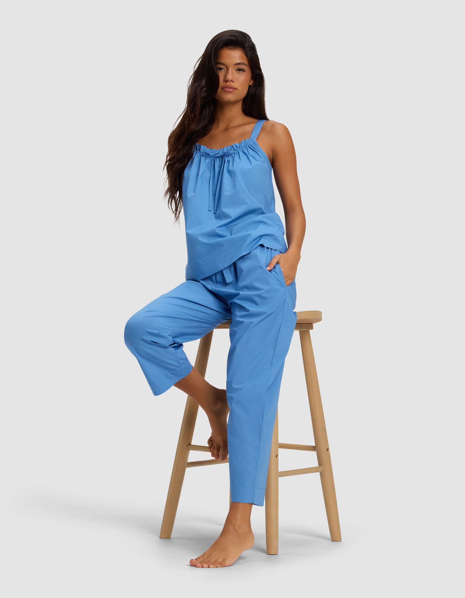 A woman with long dark hair models Cozy Earth's Women's Sutton Cropped Pajama Pant in blue, paired with a matching sleeveless top, as she sits barefoot on a wooden stool against a plain light background.