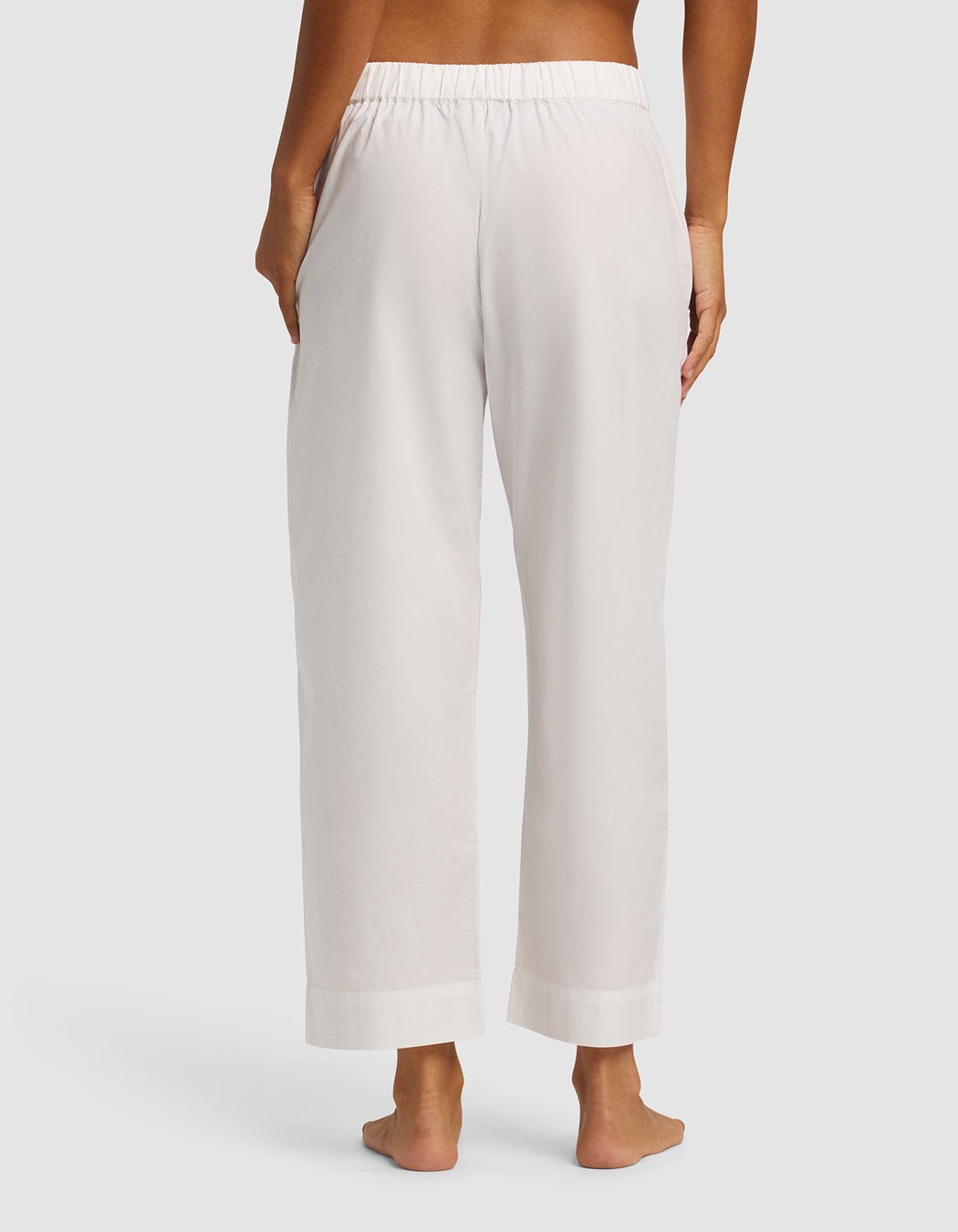 A woman is shown from the back wearing Cozy Earth's Women's Sutton Cropped Pajama Pant, featuring a loose fit, white color, elastic waistband, and side pockets; she is barefoot against a plain background.
