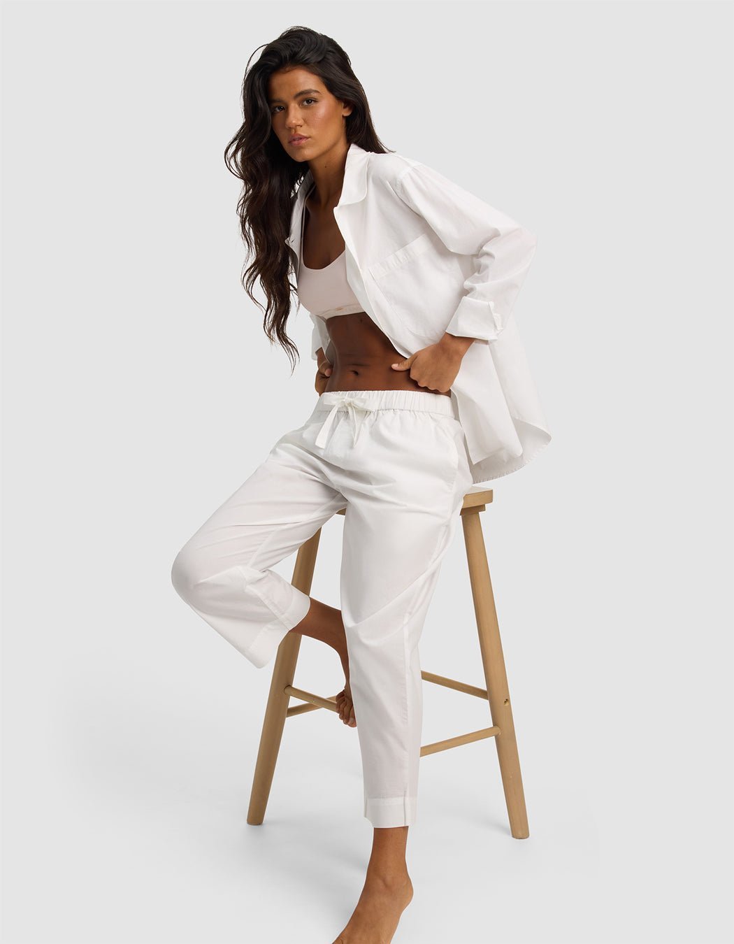 A woman with long dark hair sits on a wooden stool, wearing Cozy Earth’s Women's Sutton Cropped Pajama Pant in white with a matching loose shirt and crop top, posing relaxedly against a plain light gray background.