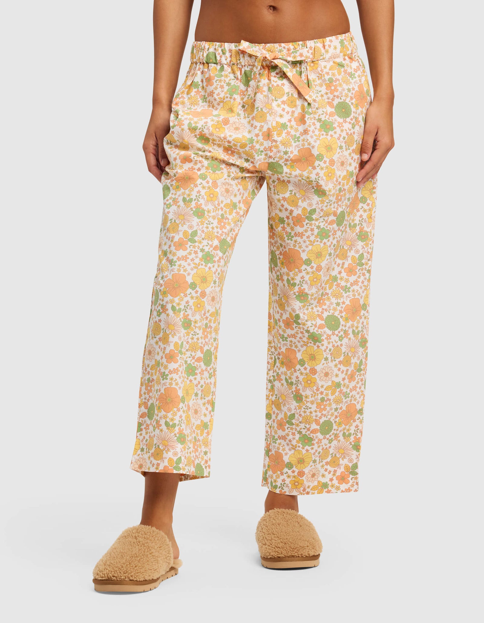 A person wears the Women's Sutton Short Sleeve & Pant Pajama Set by Cozy Earth, featuring light-colored pants with an orange, yellow, and green floral pattern, paired with beige fuzzy slippers. The image is cropped from the waist down.
