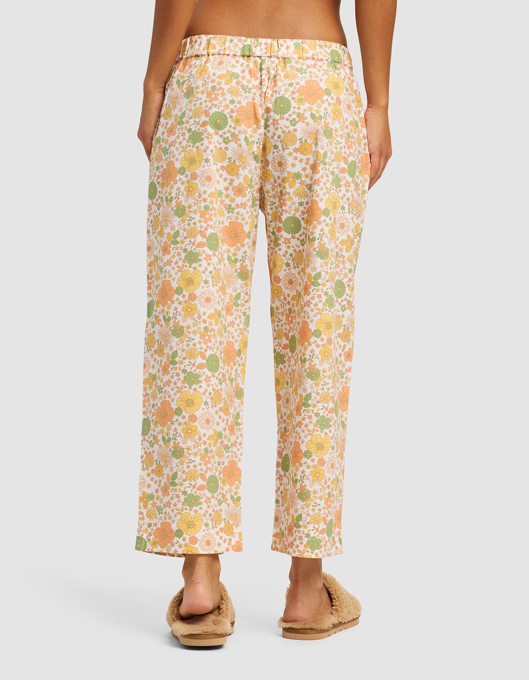 A person stands barefoot with their back to the camera, wearing Cozy Earth's Women's Sutton Cropped Pajama Pant in a loose floral print, paired with fuzzy beige slippers, against a plain background.