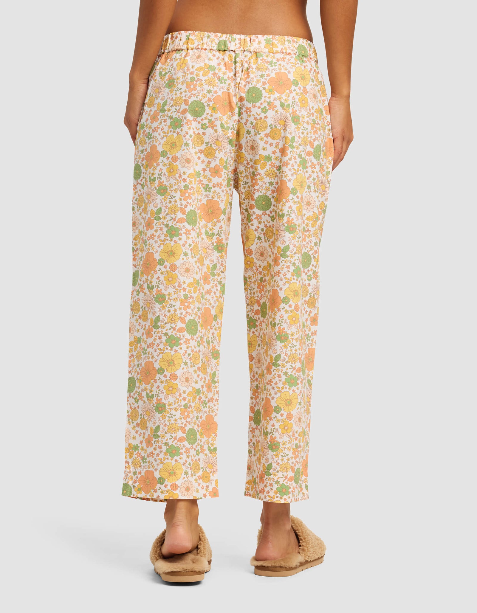 A person stands facing away, wearing Cozy Earth Women's Sutton Cropped Pajama Pant in a loose floral print with orange, yellow, and green tones, paired with beige fuzzy slippers. The relaxed-fit pants reach just above the ankles.
