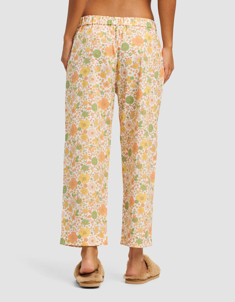A person stands facing away, wearing Cozy Earth Women's Sutton Cropped Pajama Pant in a loose floral print with orange, yellow, and green tones, paired with beige fuzzy slippers. The relaxed-fit pants reach just above the ankles.