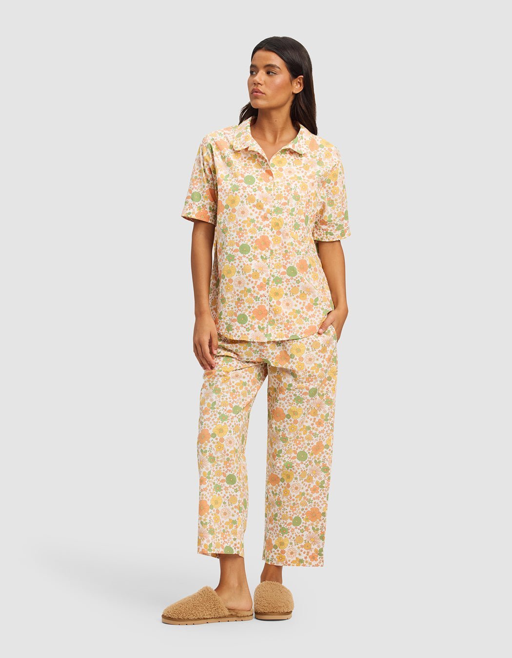 A woman models the Cozy Earth Women's Sutton Cropped Pajama Pant with a matching pastel floral short-sleeve button-up top and tan fuzzy slippers, standing against a plain light gray background.