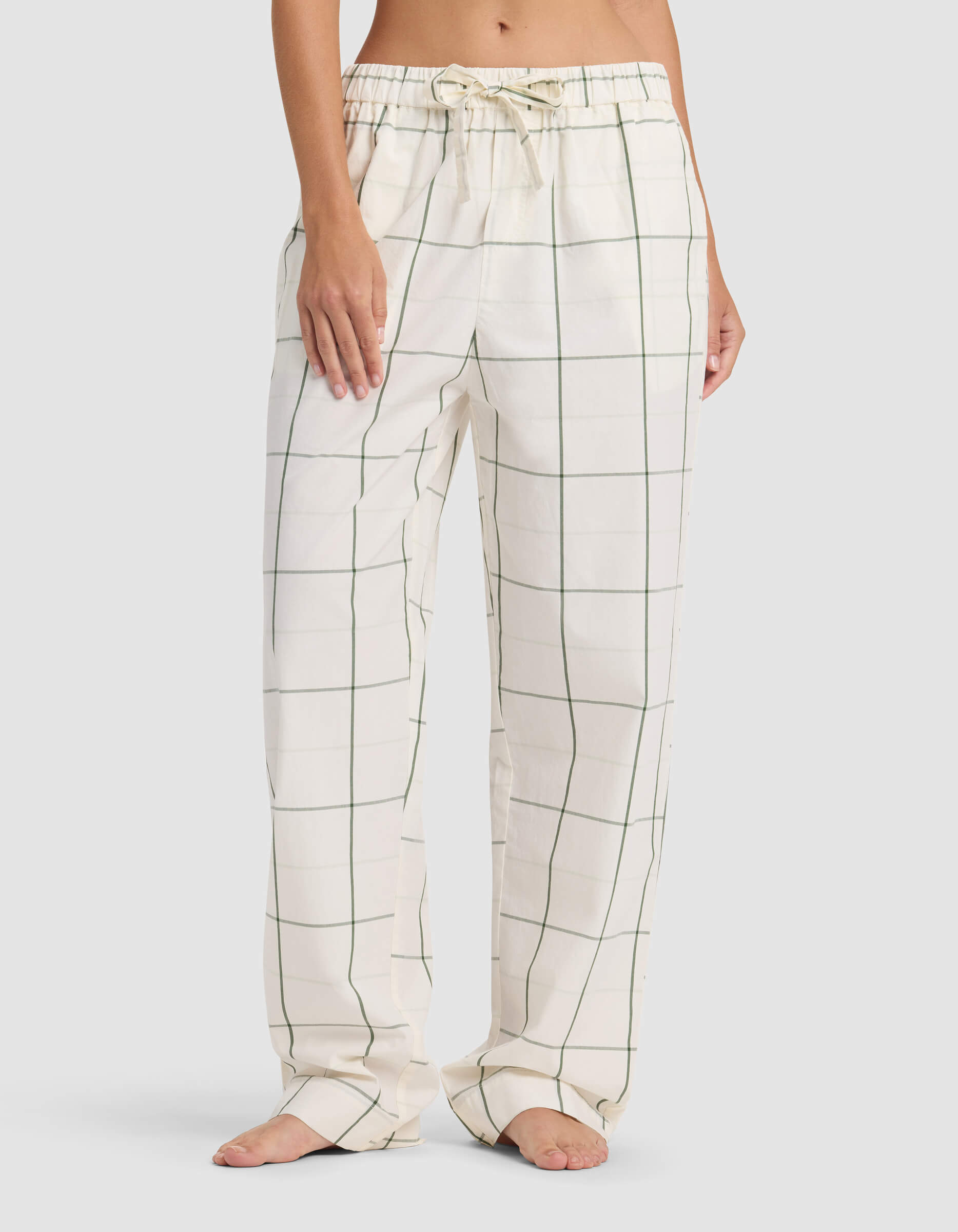 A person wears Cozy Earth Women's Sutton Pajama Pant—loose-fitting cream with a green windowpane check and an adjustable drawstring waist—standing barefoot. Their upper body is mostly out of the frame.