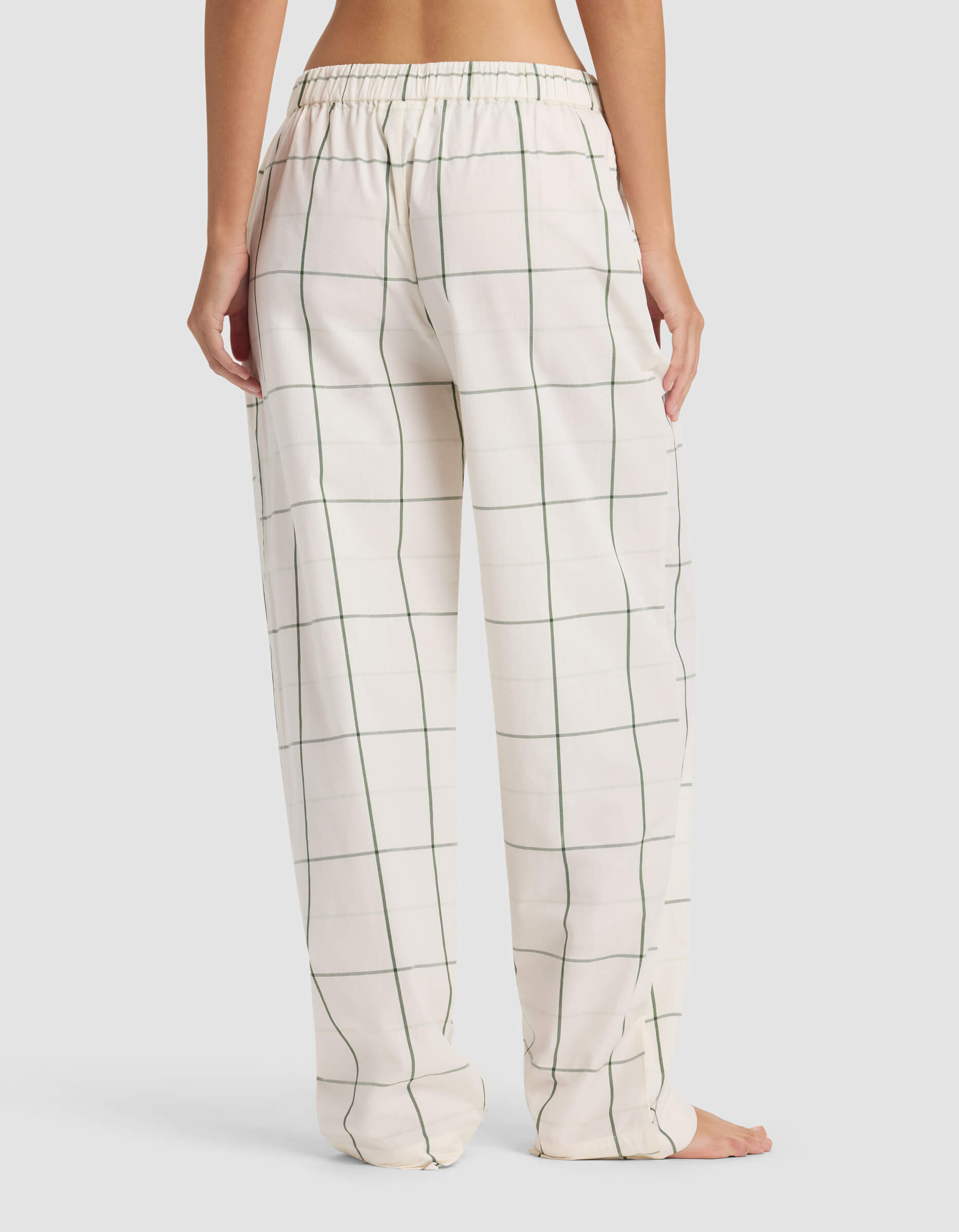 A woman stands barefoot, shown from the back, wearing Cozy Earth Women's Sutton Pajama Pant—loose-fitting white pants with a green windowpane check pattern and an elastic waistband.