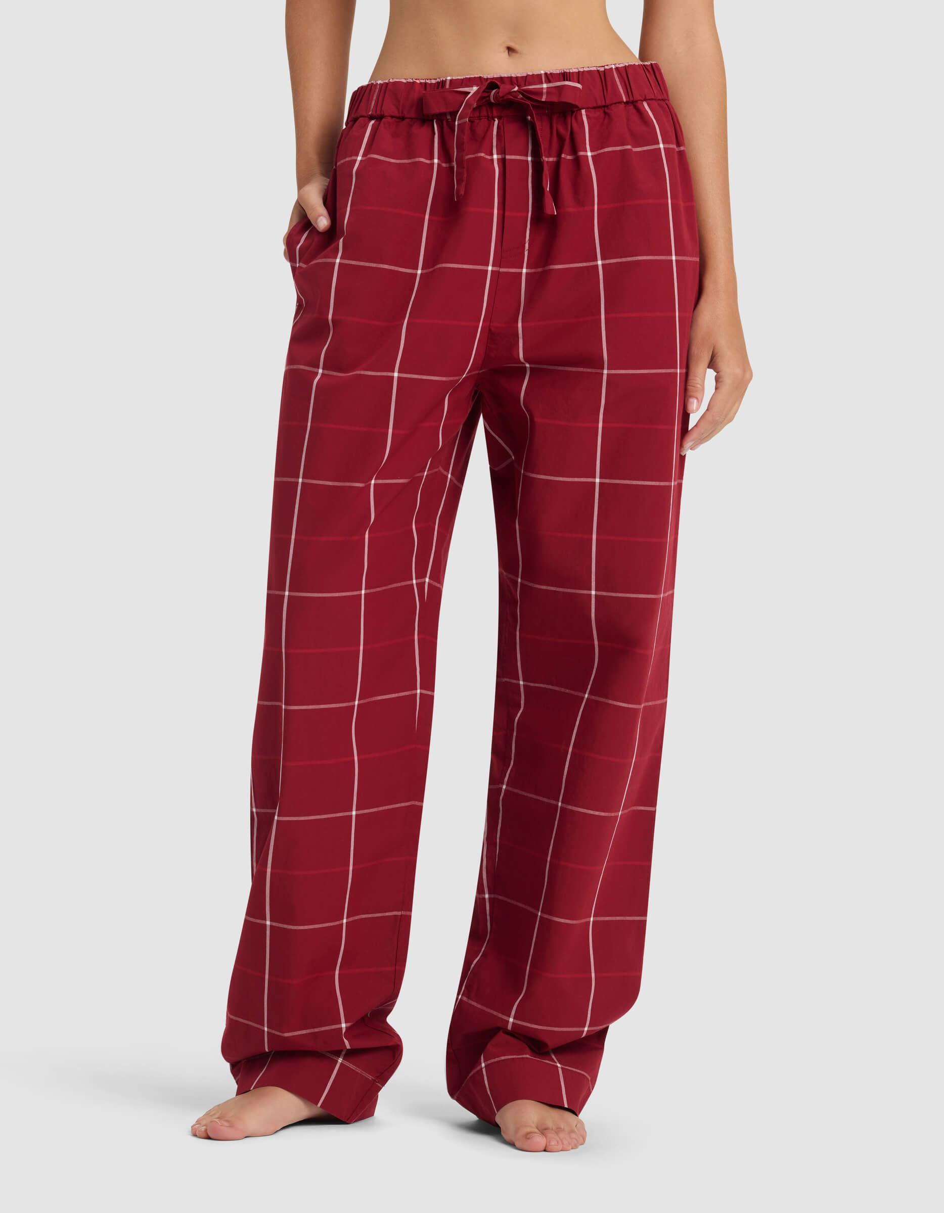 A woman wearing Cozy Earth’s Women’s Sutton Pajama Pant—loose-fitting red pants with a white windowpane check, drawstring waist, and side pockets—stands barefoot against a plain background.|Color:Garnet Windowpane