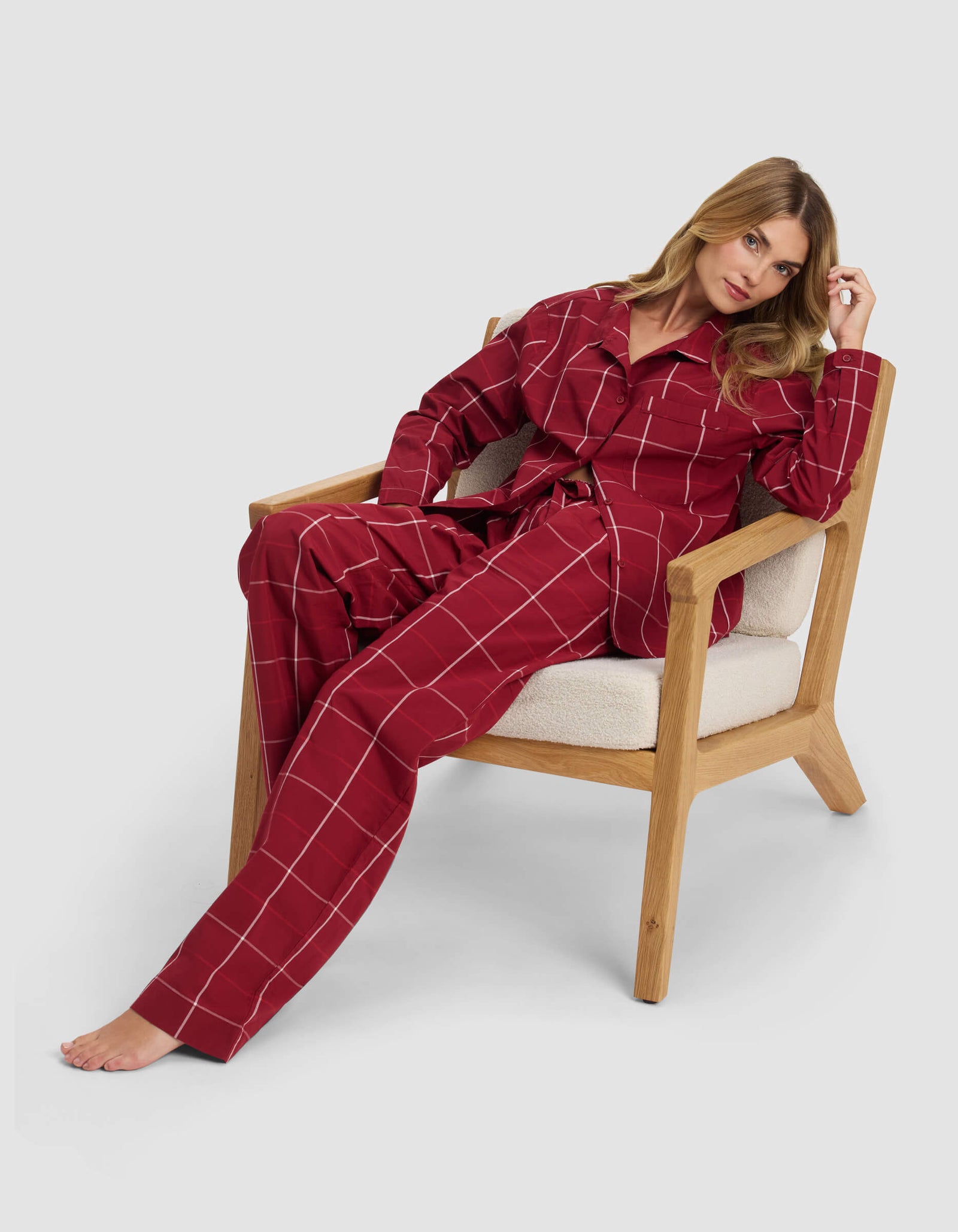 A woman sits barefoot on a wooden chair, wearing the Cozy Earth Women's Sutton Pajama Pant in red with white grid lines. She leans back, one arm on the armrest, and tilts her head slightly to the side.