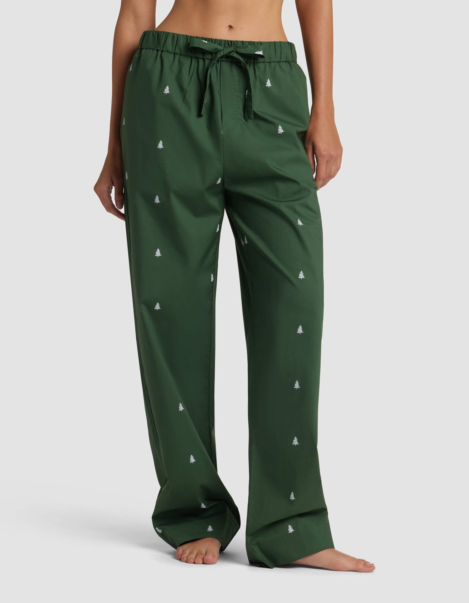 A person stands barefoot against a plain background wearing Cozy Earth’s Women’s Sutton Pajama Pant in dark green, featuring a drawstring waist and small white tree patterns. The loose-fitting pants reveal the person’s midriff and feet.