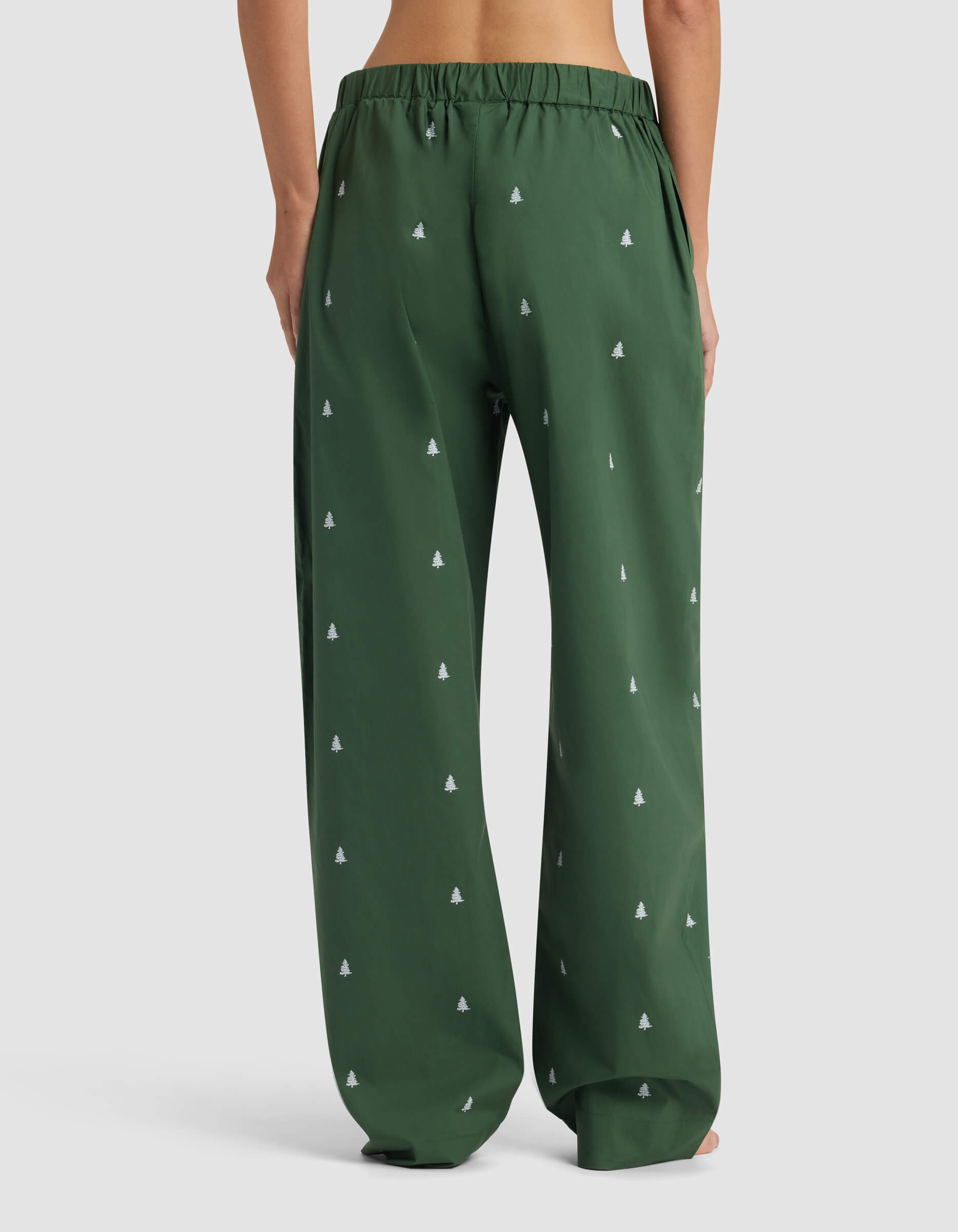 A woman models the Women's Sutton Pajama Pant by Cozy Earth, shown from the back waist down in loose-fitting dark green fabric with a subtle white tree pattern against a plain light background.