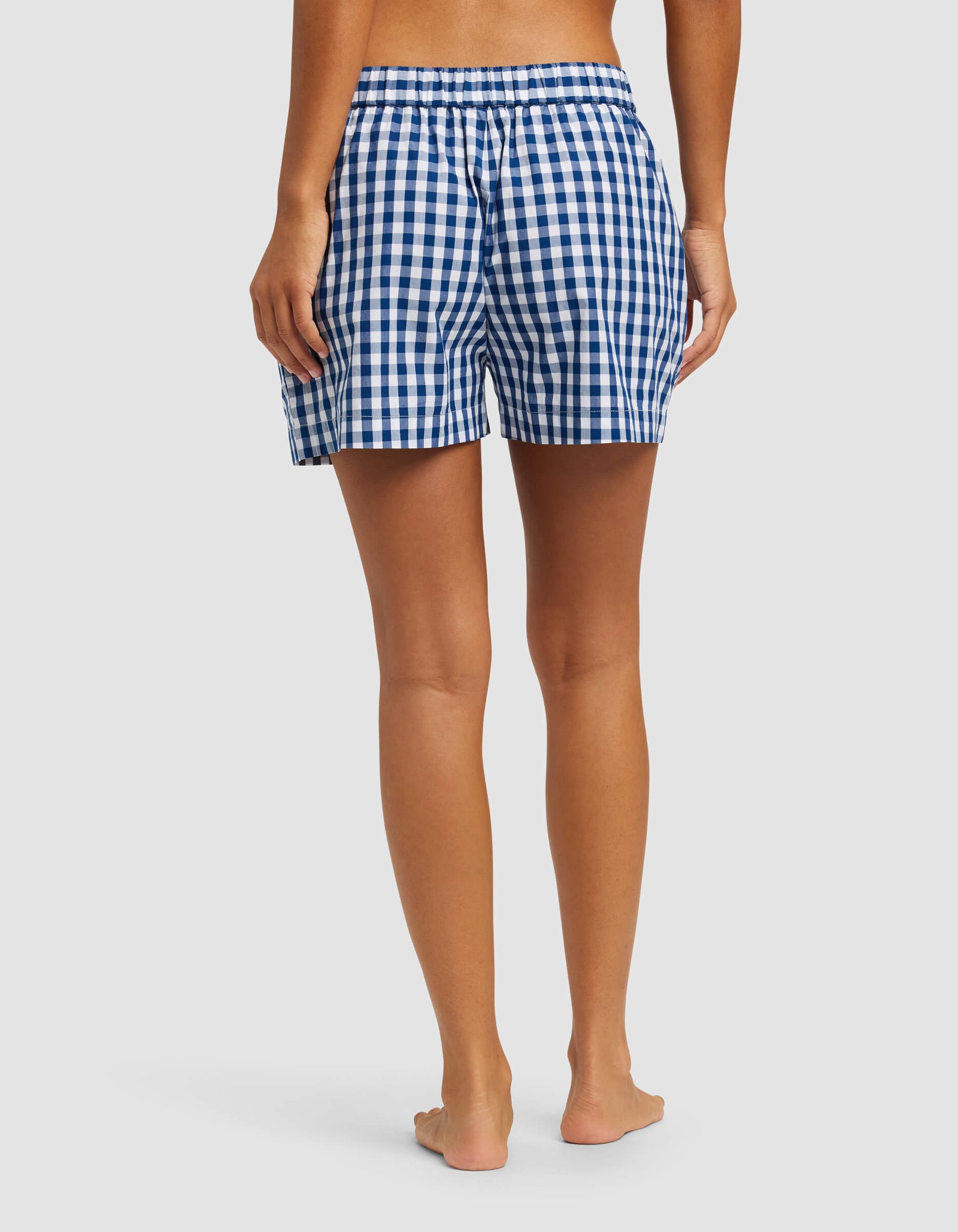 A person stands barefoot facing away, wearing Cozy Earth's Women's Sutton Pajama Short—blue and white checkered with an elastic waistband—against a plain light background.