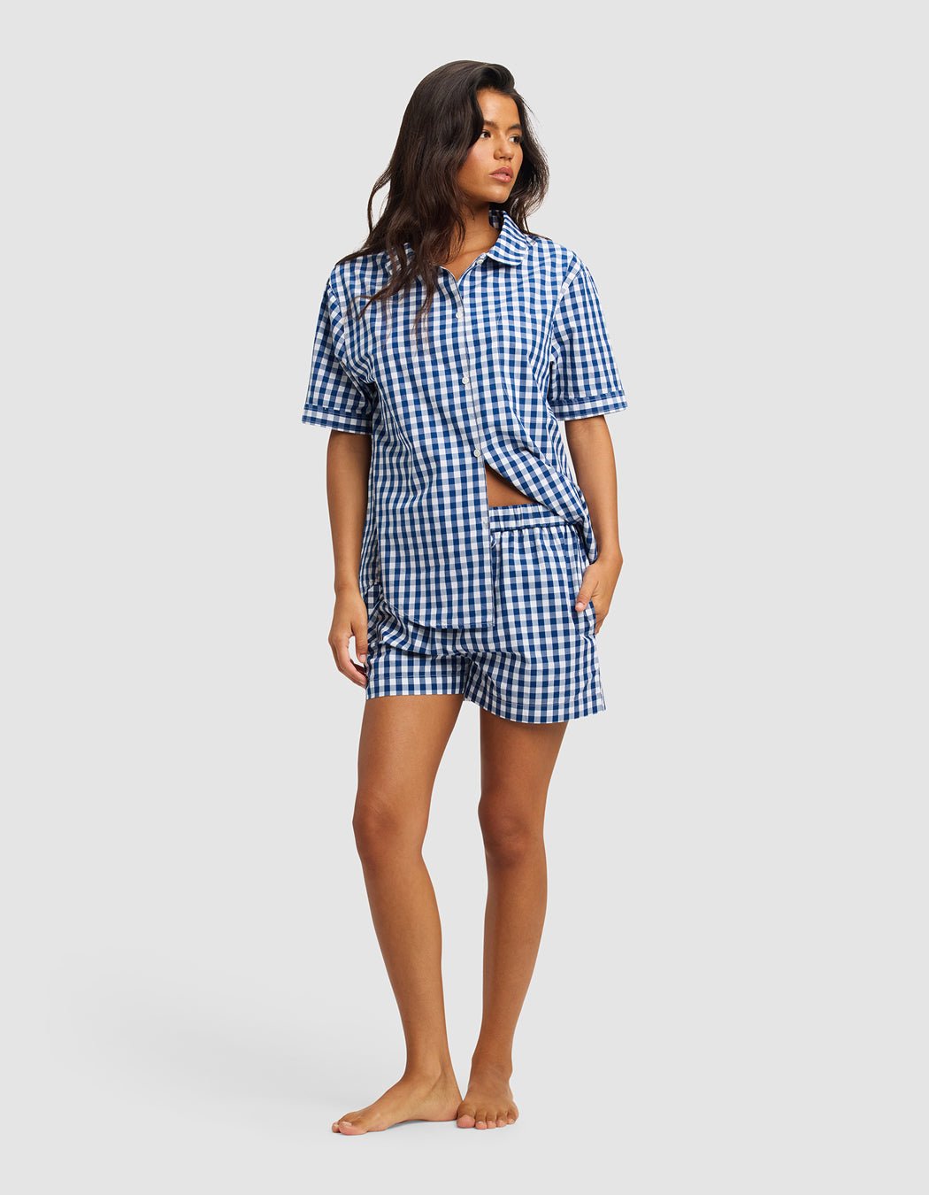 A woman stands barefoot wearing Cozy Earth's Women's Sutton Pajama Short, a blue-and-white gingham short-sleeve set with matching shorts and a button-up shirt, against a plain light gray background.