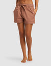 A woman stands barefoot with hands in pockets, wearing Cozy Earth's Women's Sutton Pajama Short in light brown. The photo, cropped from the waist down, is set against a plain light background.|Color:Saddle