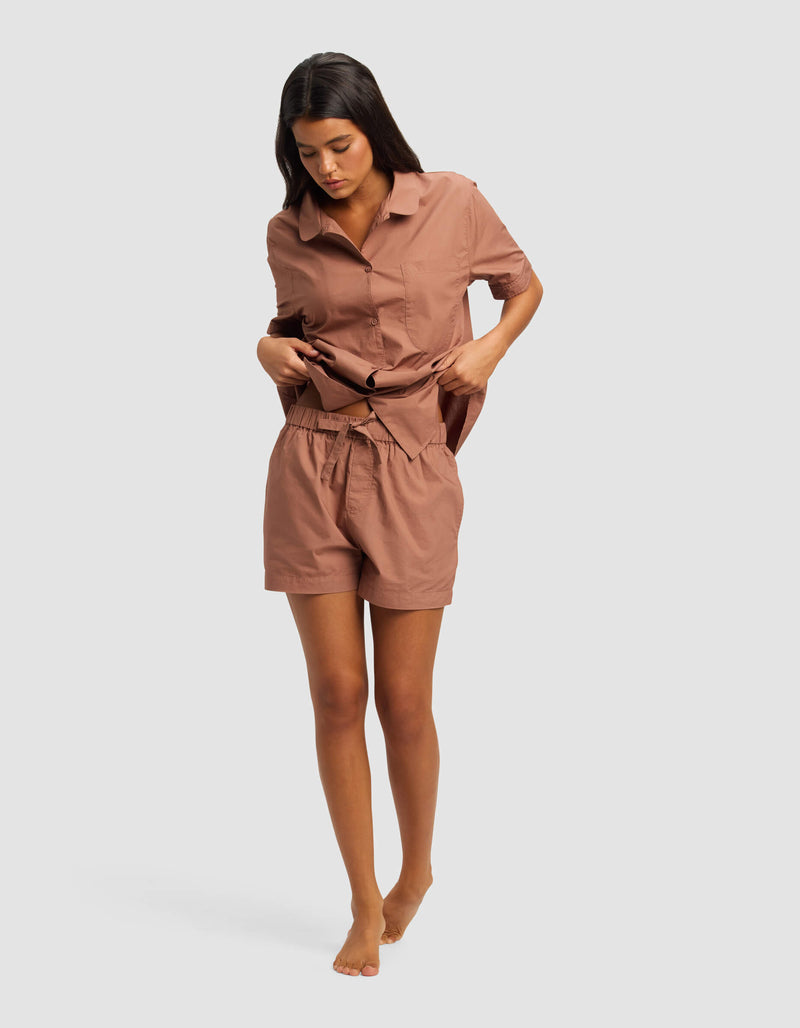 A woman wearing Cozy Earth's Women's Sutton Pajama Short with a matching loose, short-sleeved, button-up shirt stands barefoot against a plain light background, looking down and adjusting her shirt.