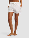 A person stands barefoot with hands in pockets, modeling the Cozy Earth Women's Sutton Pajama Short in white against a plain light gray background. The upper body is not shown.|Color:White