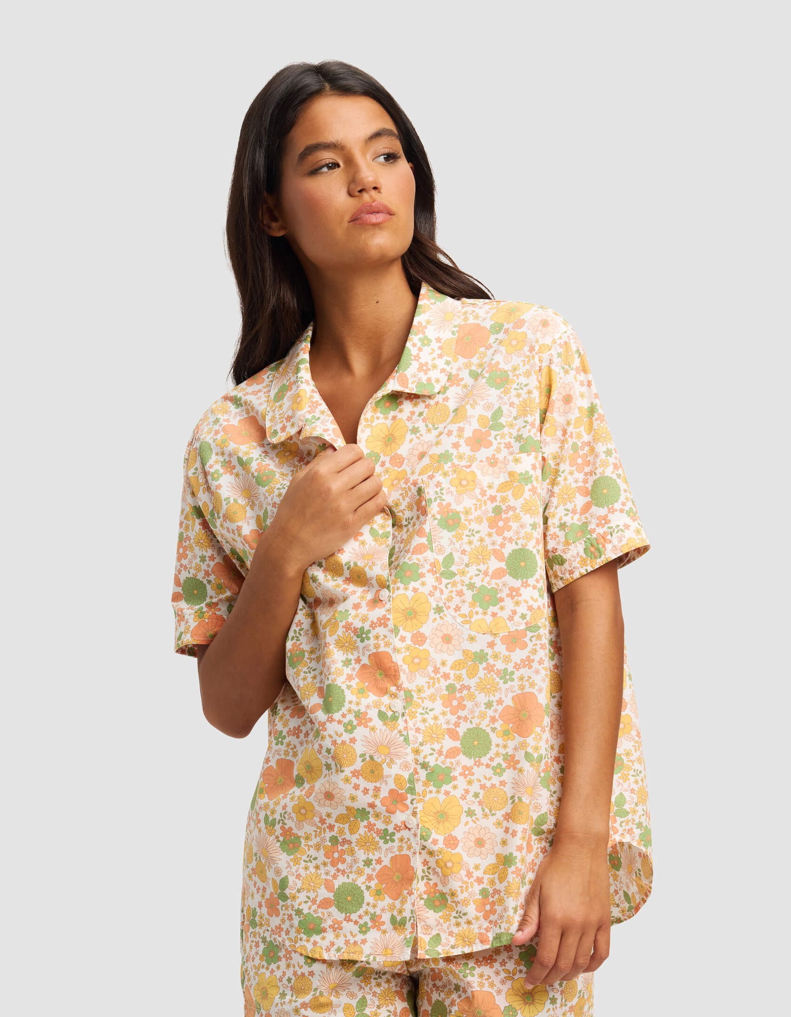 A woman wears the Cozy Earth Women's Sutton Pajama Short set in a pastel floral pattern, standing against a light gray background and looking to the side with a relaxed expression.