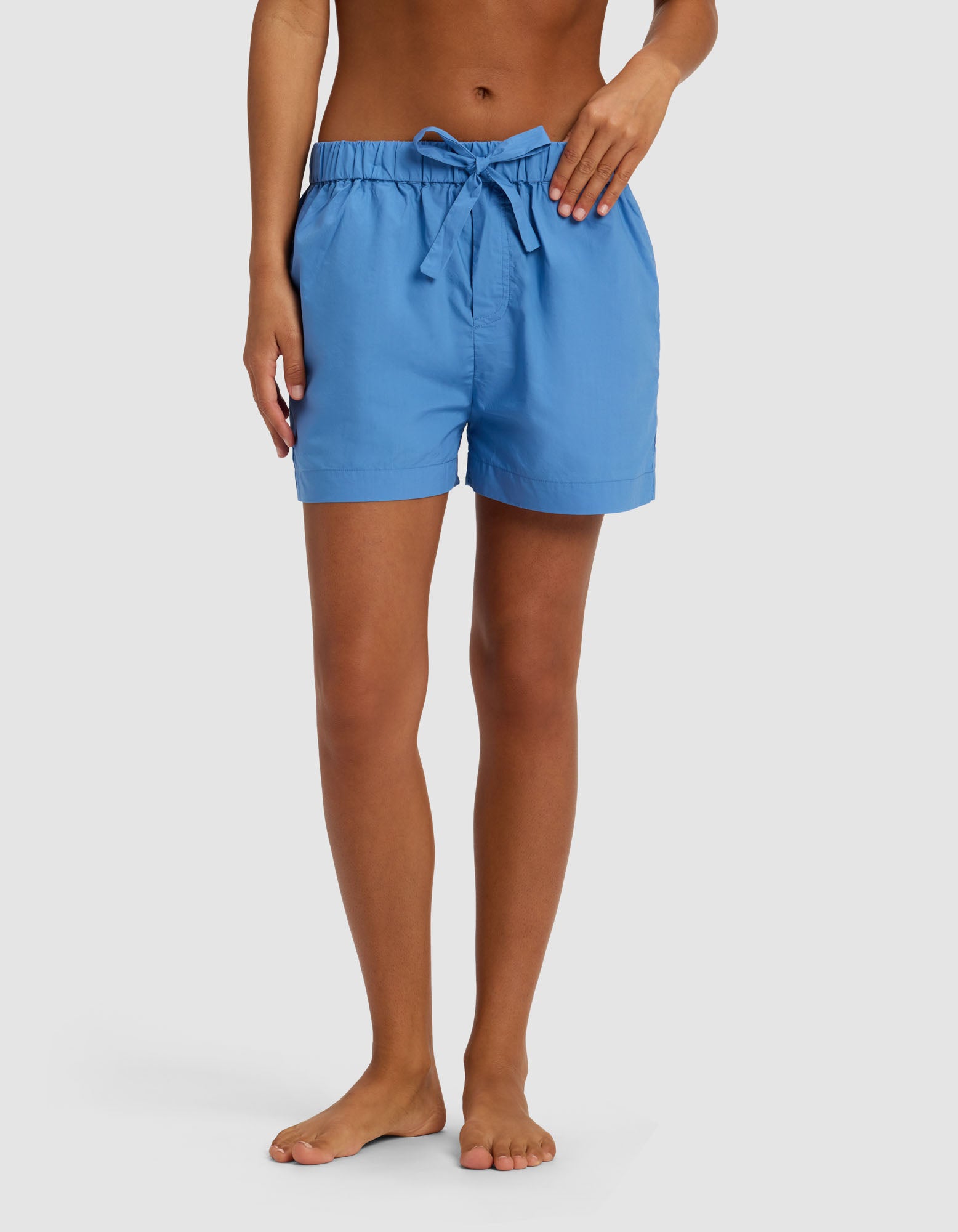 A person stands barefoot against a plain white background, wearing Cozy Earth's Women's Sutton Pajama Short in light blue with a tied drawstring bow at the waist.|Color:Sailor