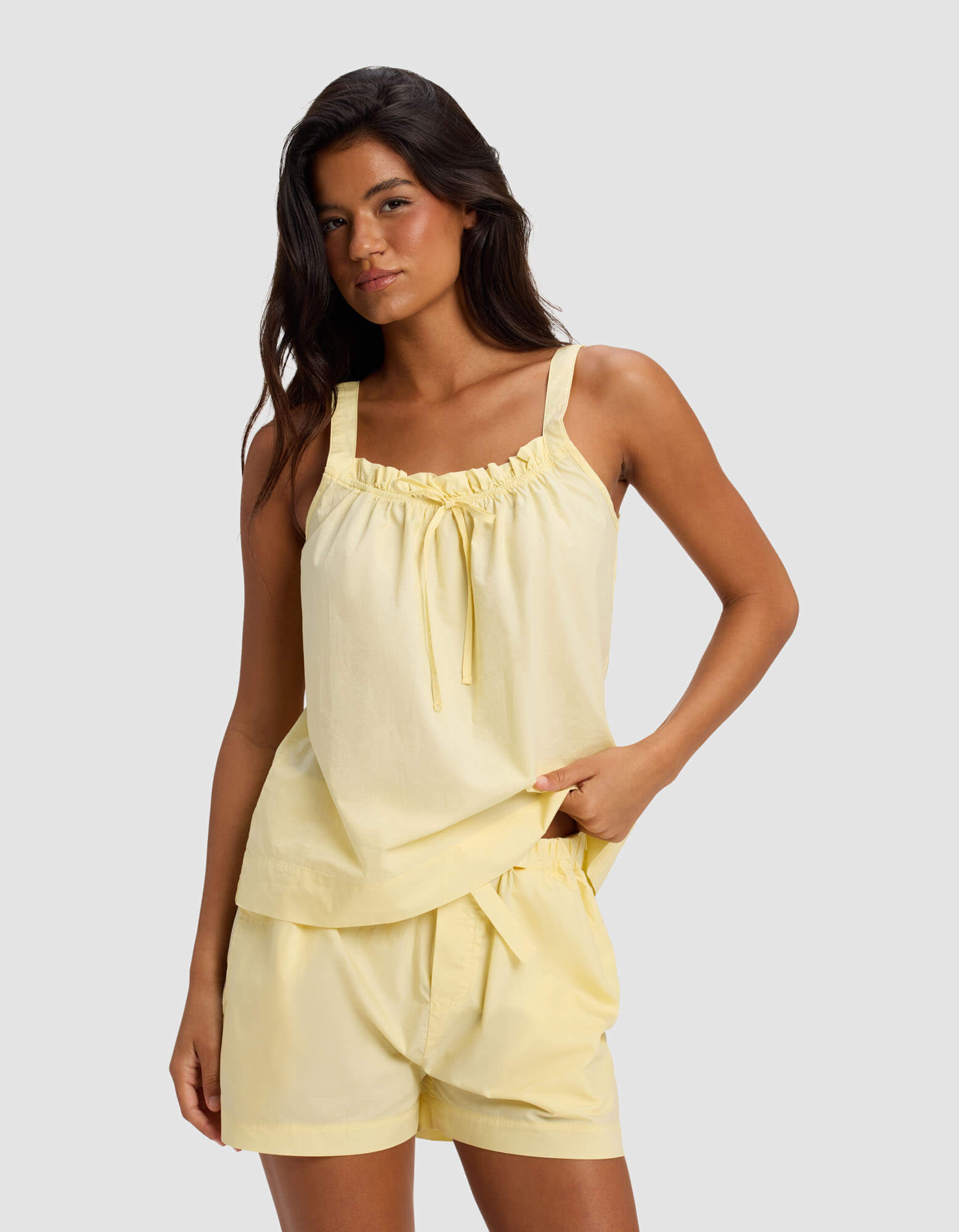 A woman wears Cozy Earth's Women's Sutton Pajama Tank in Meringue, featuring a tie neckline, paired with matching loose shorts, standing against a plain light gray background.