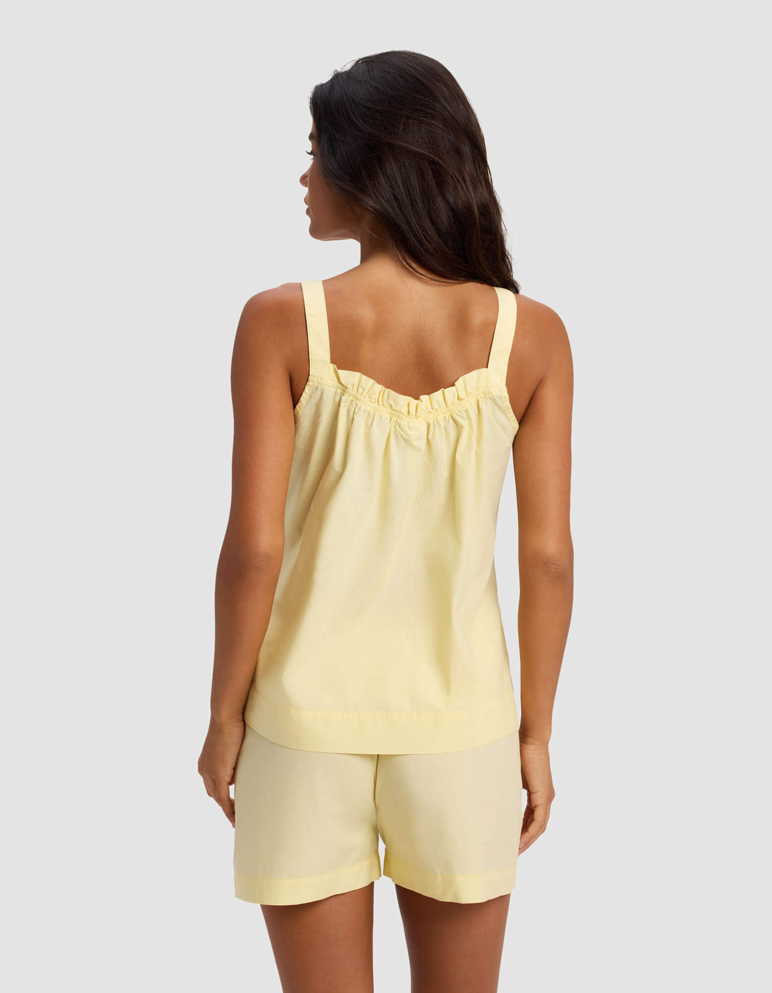 A woman with long dark hair, shown from the back, wears the Cozy Earth Women's Sutton Pajama Tank in light yellow with ruffle detail and matching shorts, standing against a plain light gray background.|Color:Meringue
