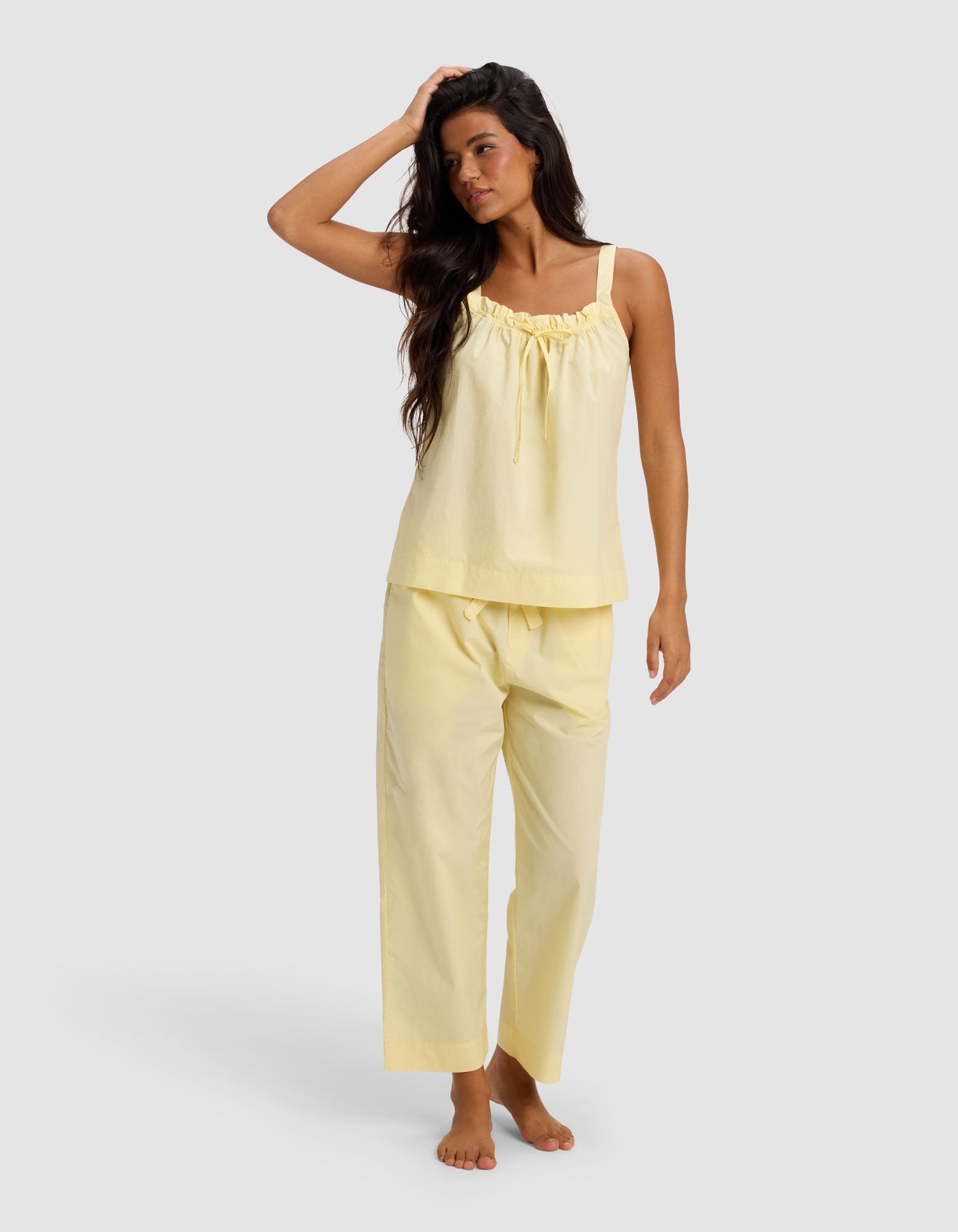 A woman with long dark hair poses barefoot in Cozy Earth's Women's Sutton Tank & Cropped Pant Pajama Set—light yellow, sleeveless with ruffle detail and matching loose pants—against a plain light gray background.|Color:Meringue