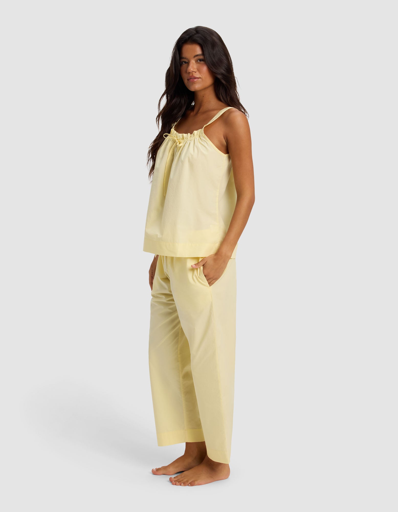 A woman stands barefoot on a plain background wearing the Cozy Earth Women's Sutton Tank & Cropped Pant Pajama Set in light yellow, featuring a loose sleeveless top with ruffle detail and matching wide-leg pants. Her long hair is down, hands in pockets.