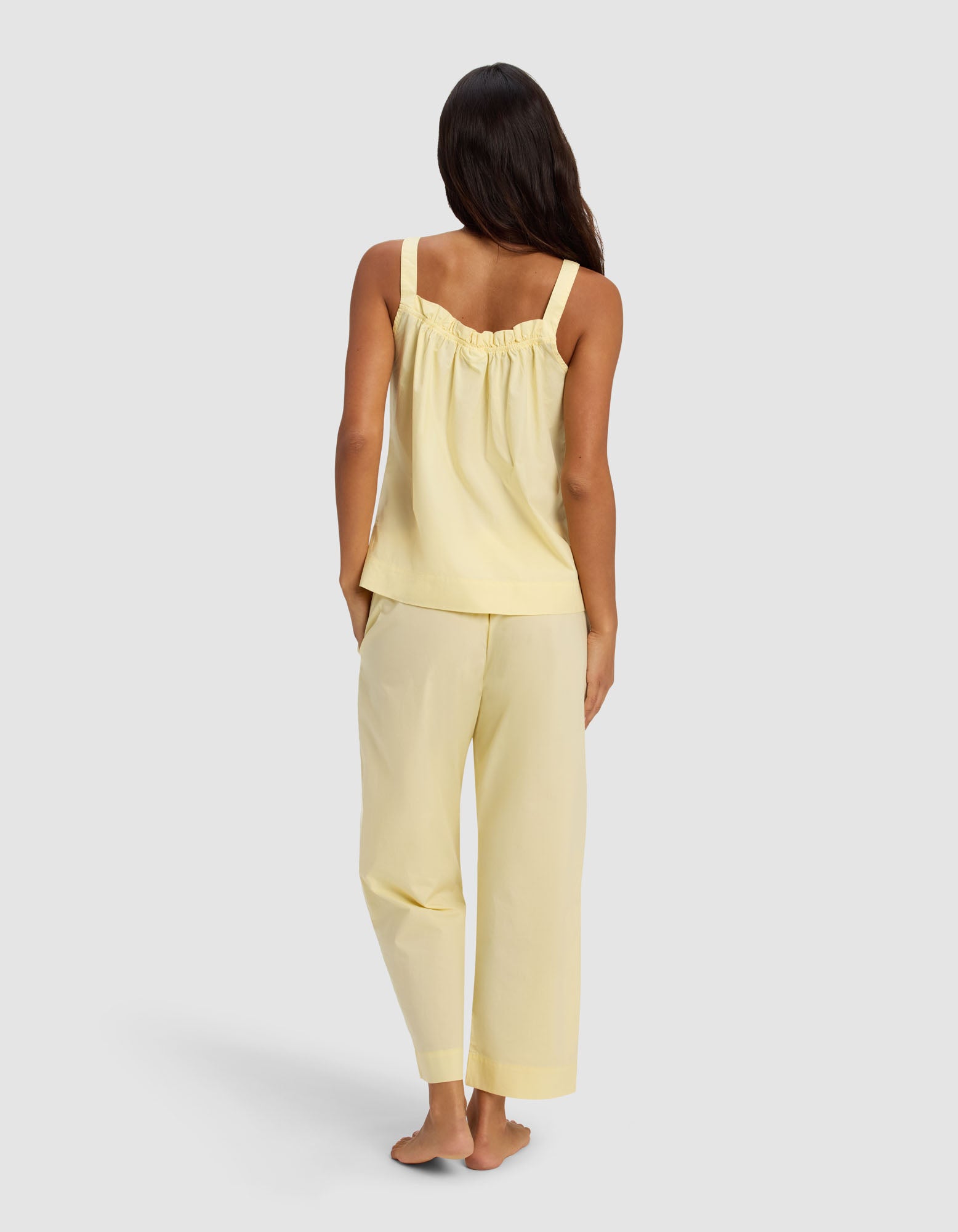 A woman with long dark hair stands barefoot, facing away, wearing the Cozy Earth Women's Sutton Tank & Cropped Pant Pajama Set in yellow against a plain light gray background.