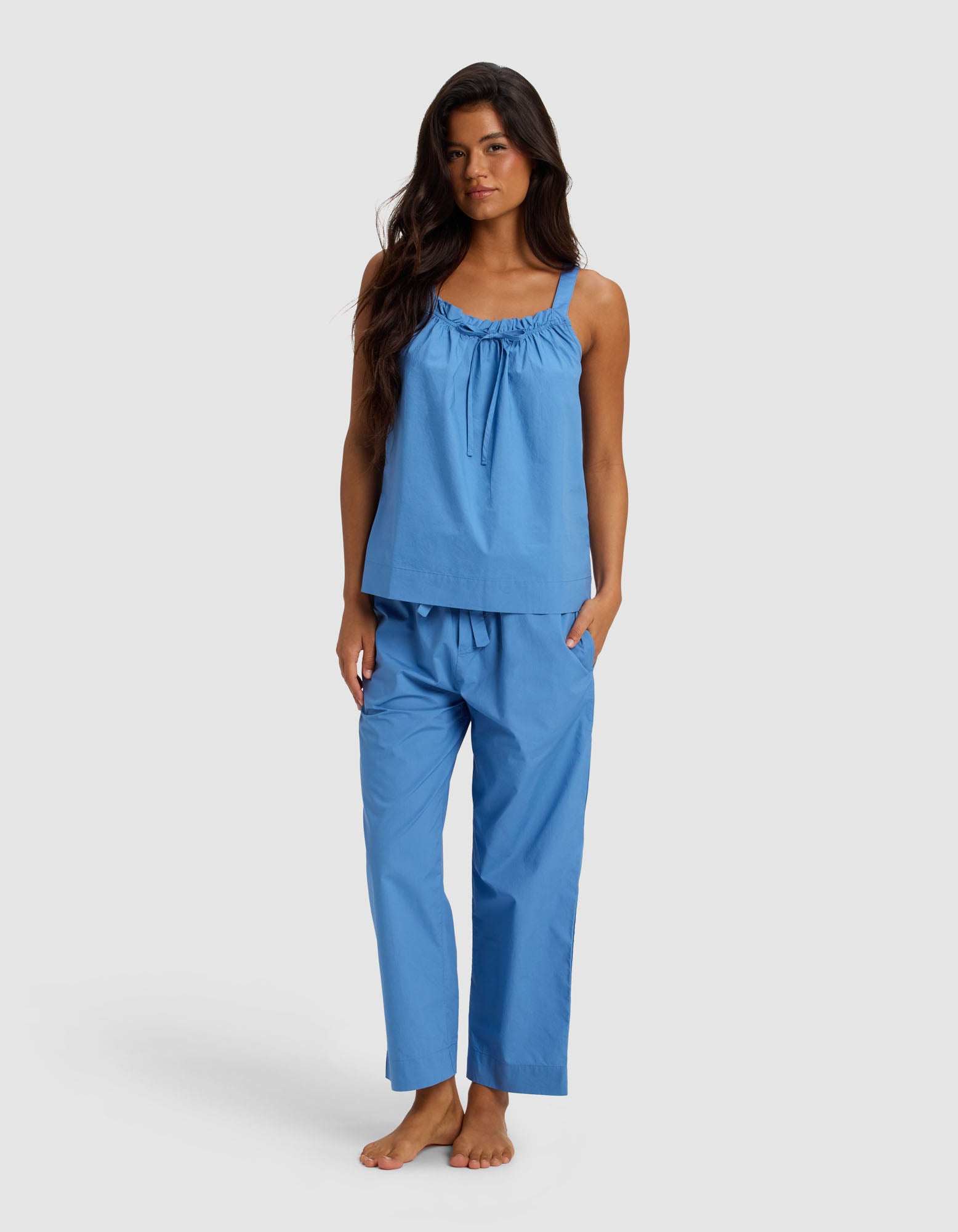 A woman with long dark hair stands barefoot against a plain light background, wearing the Cozy Earth Women's Sutton Tank & Cropped Pant Pajama Set in light blue.