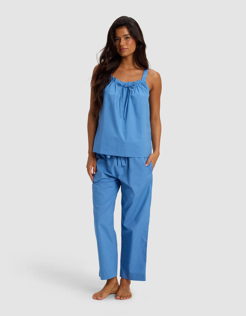 A woman with long dark hair stands barefoot against a plain light background, wearing the Cozy Earth Women's Sutton Tank & Cropped Pant Pajama Set in light blue.