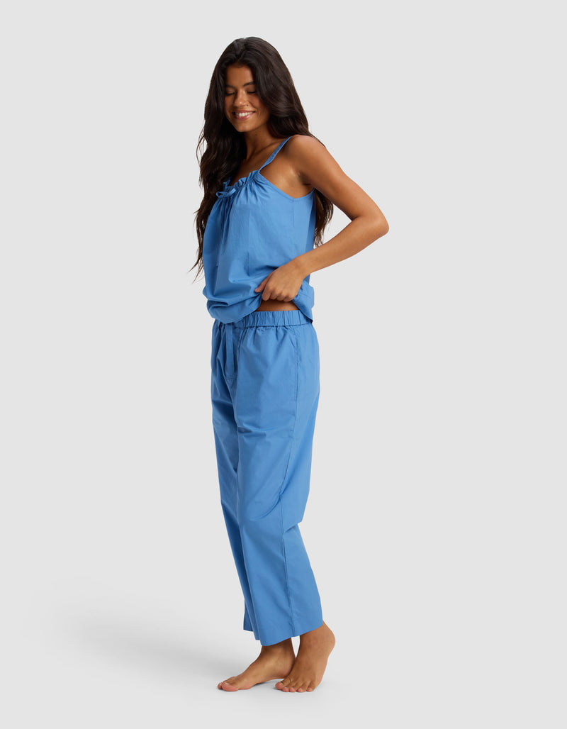 A woman with long dark hair stands barefoot, smiling in the Cozy Earth Women's Sutton Tank & Cropped Pant Pajama Set—a loose-fitting blue sleeveless top with thin straps and matching pants—against a plain light background.