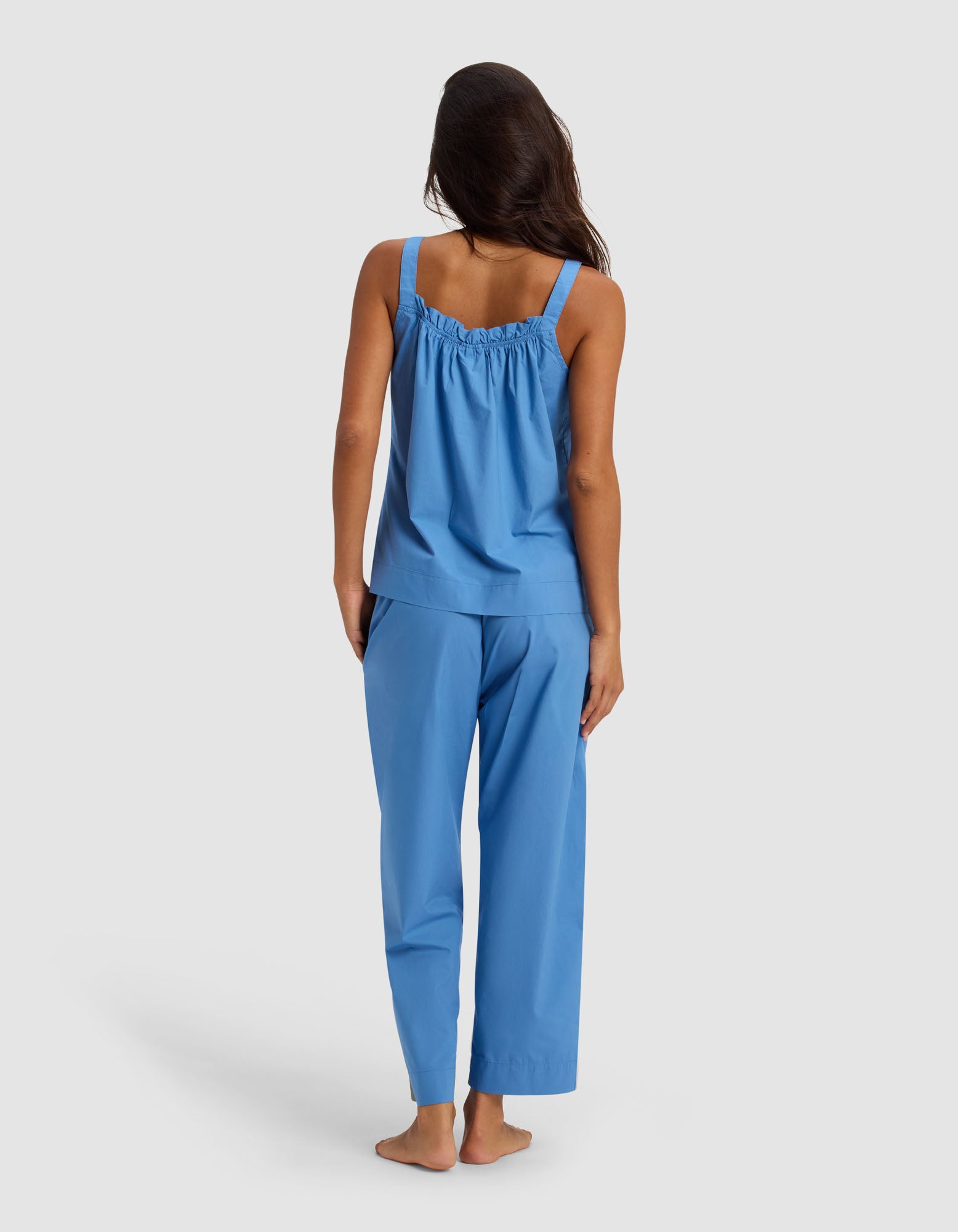 A woman stands barefoot facing away, wearing the Cozy Earth Women's Sutton Tank & Cropped Pant Pajama Set in blue against a plain, light background.