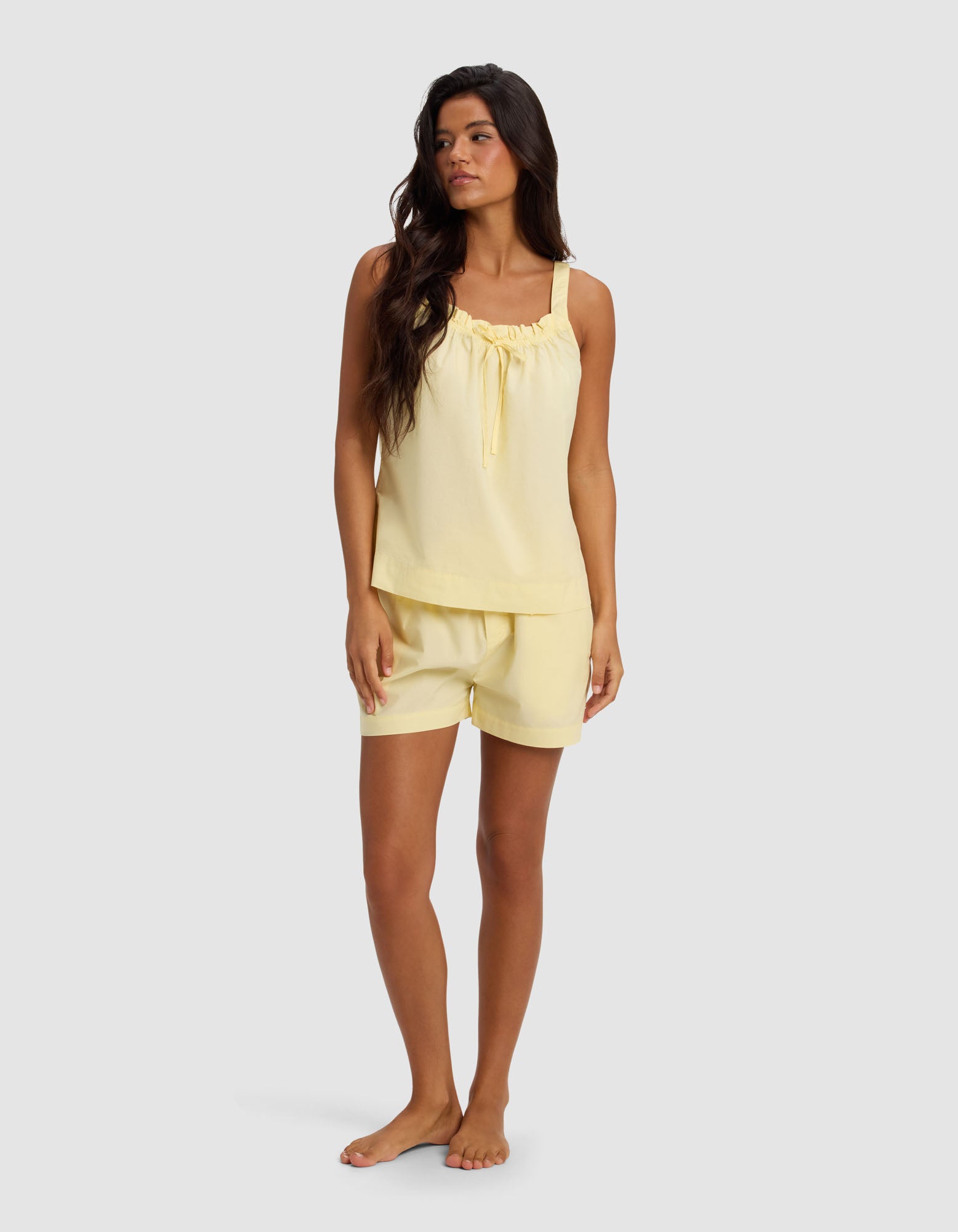 A woman with long dark hair wears the Cozy Earth Women's Sutton Tank & Short Pajama Set in light yellow, featuring a sleeveless top with a gathered neckline and matching shorts, standing barefoot against a plain light gray background.