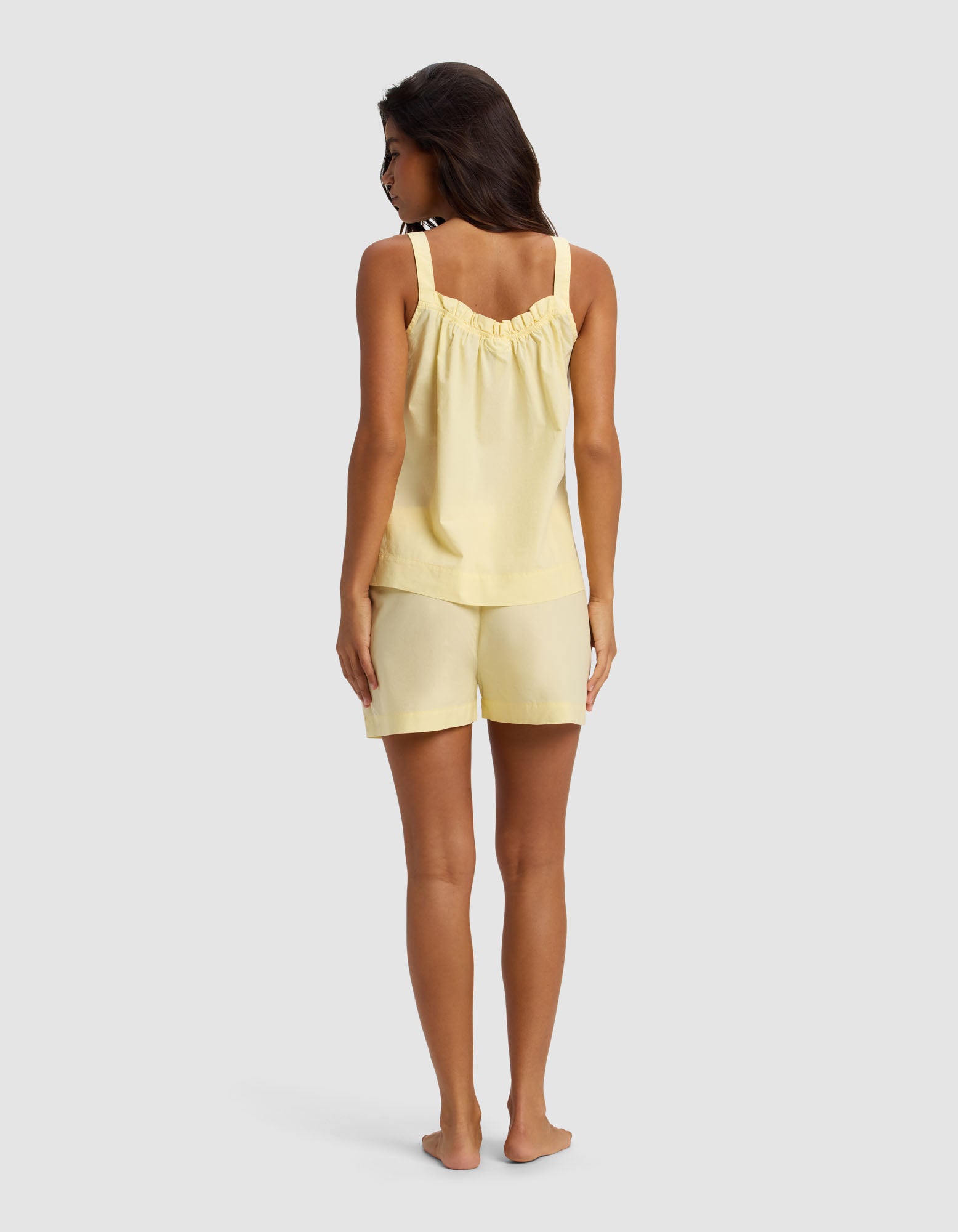 A woman stands barefoot facing away against a light gray background, wearing the Cozy Earth Women's Sutton Tank & Short Pajama Set in pale yellow with ruffled straps.