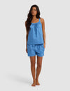 A woman with long, wavy dark hair stands barefoot against a light gray background, wearing the Cozy Earth Women's Sutton Tank & Short Pajama Set—an easygoing, sleeveless blue top with a gathered neckline and matching shorts.|Color:Sailor