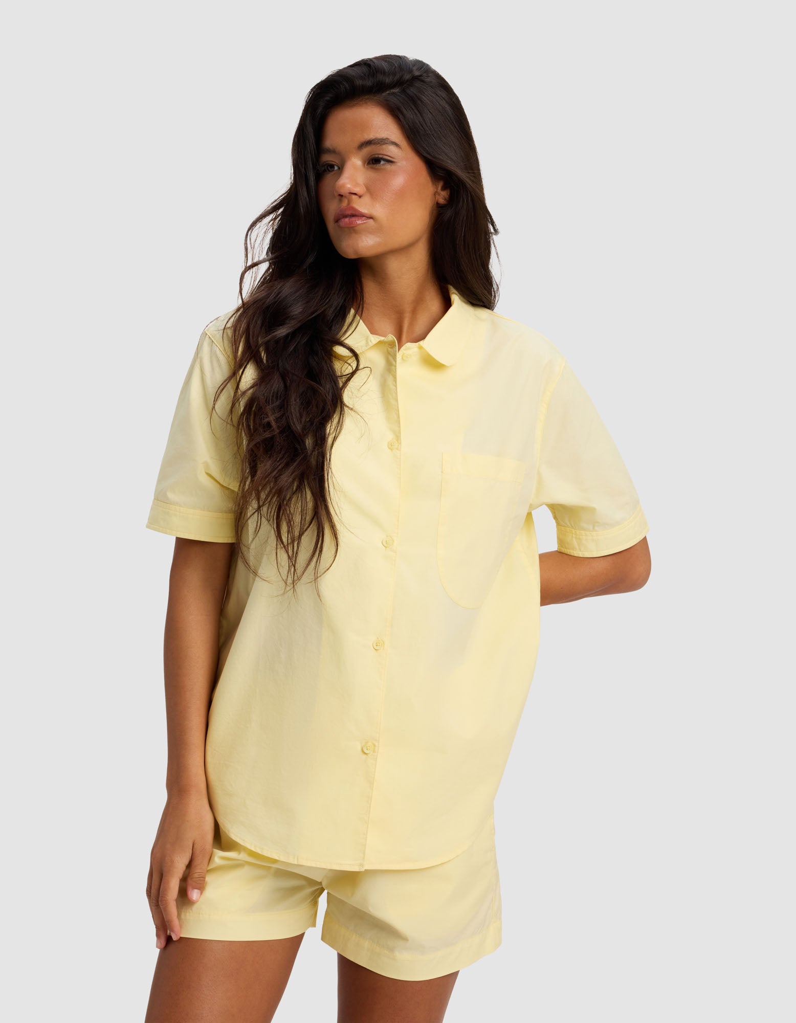 A woman with long dark hair wears the Cozy Earth Women's Sutton Short Sleeve Pajama Top in light yellow, paired with matching shorts, standing against a plain light gray background.