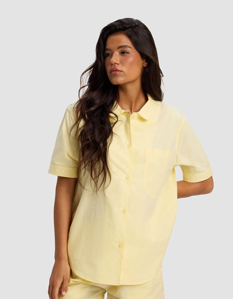 A woman with long, wavy dark hair models the Cozy Earth Women's Sutton Short Sleeve & Cropped Pant Pajama Set in light yellow, standing against a plain light gray background and looking to her left.