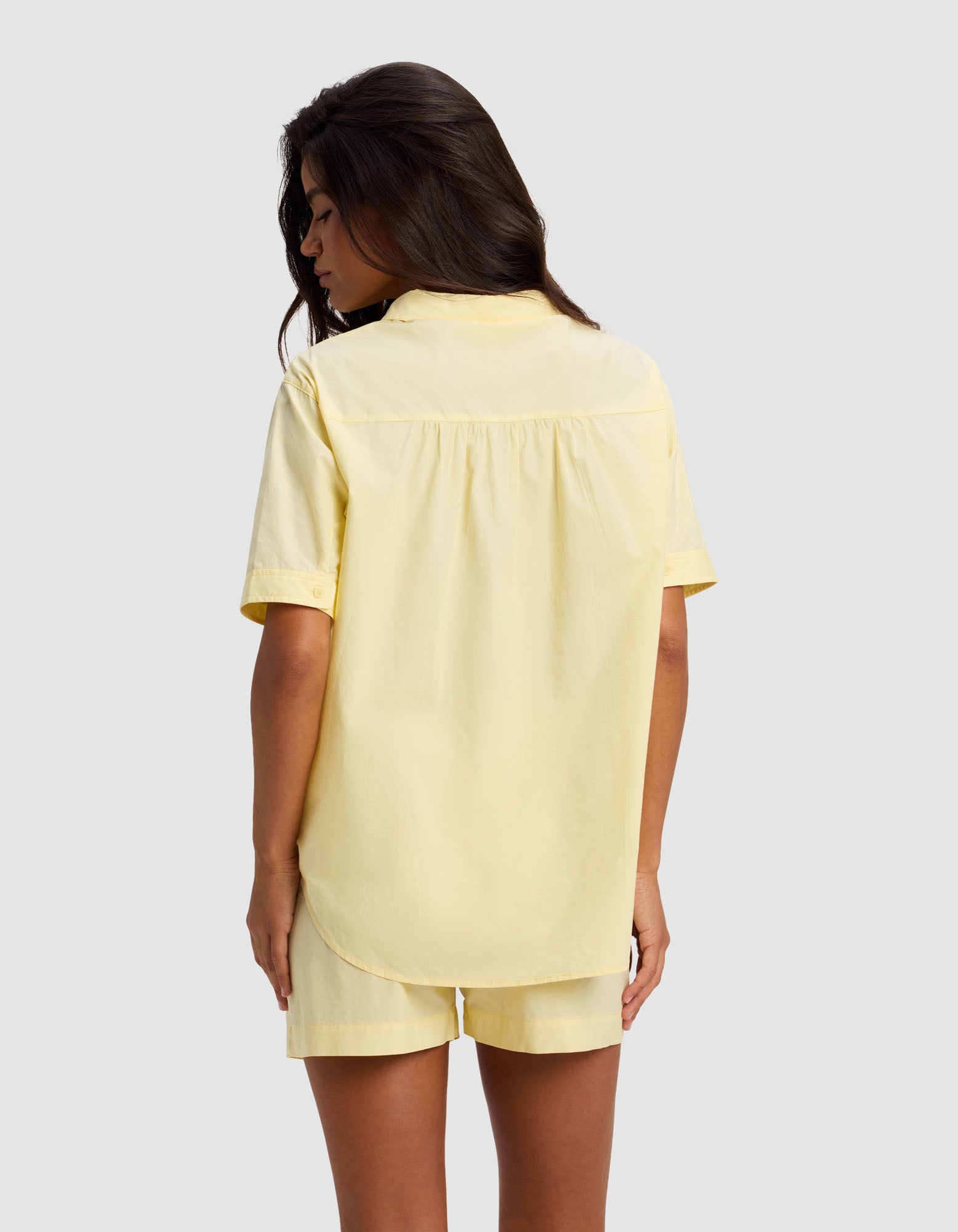 A woman with long dark hair is seen from behind, wearing the Cozy Earth Women's Sutton Short Sleeve Pajama Top in pale yellow, paired with matching shorts, standing against a light gray background.