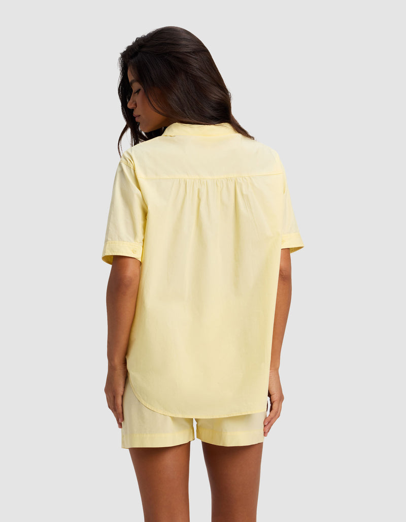 A woman with long dark hair is seen from behind, wearing the Cozy Earth Women's Sutton Short Sleeve Pajama Top in pale yellow, paired with matching shorts, standing against a light gray background.