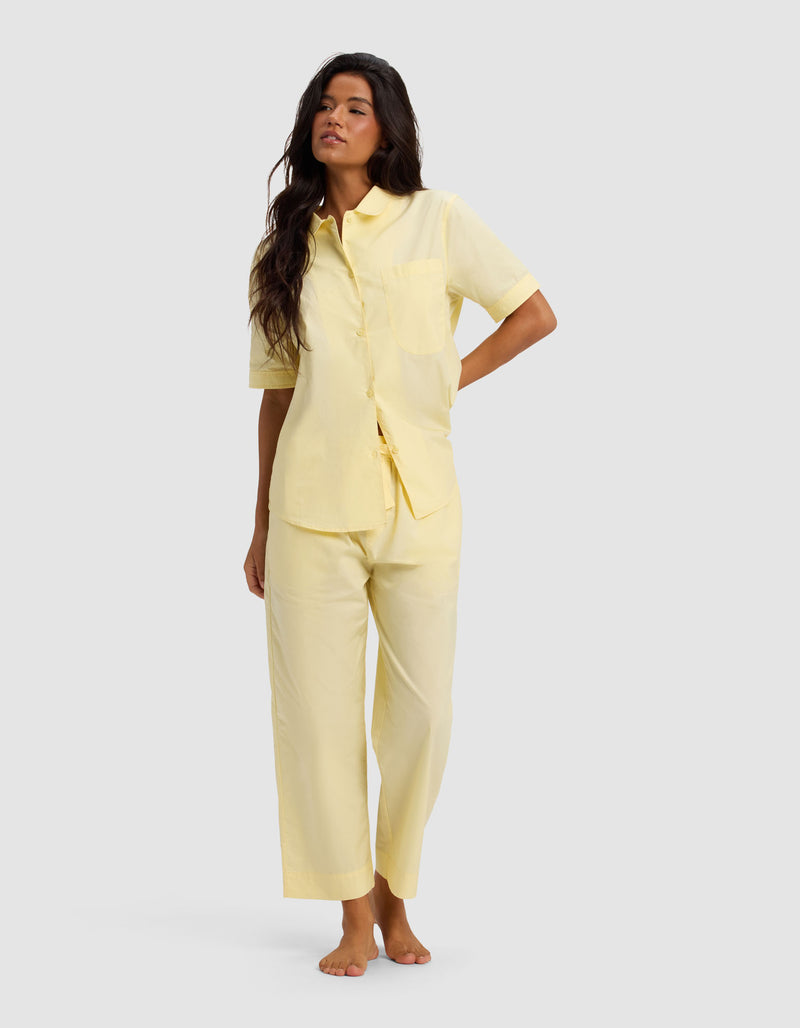 A woman with long, wavy dark hair stands barefoot against a light gray background, wearing the Cozy Earth Women's Sutton Short Sleeve & Cropped Pant Pajama Set in light yellow. She looks relaxed.