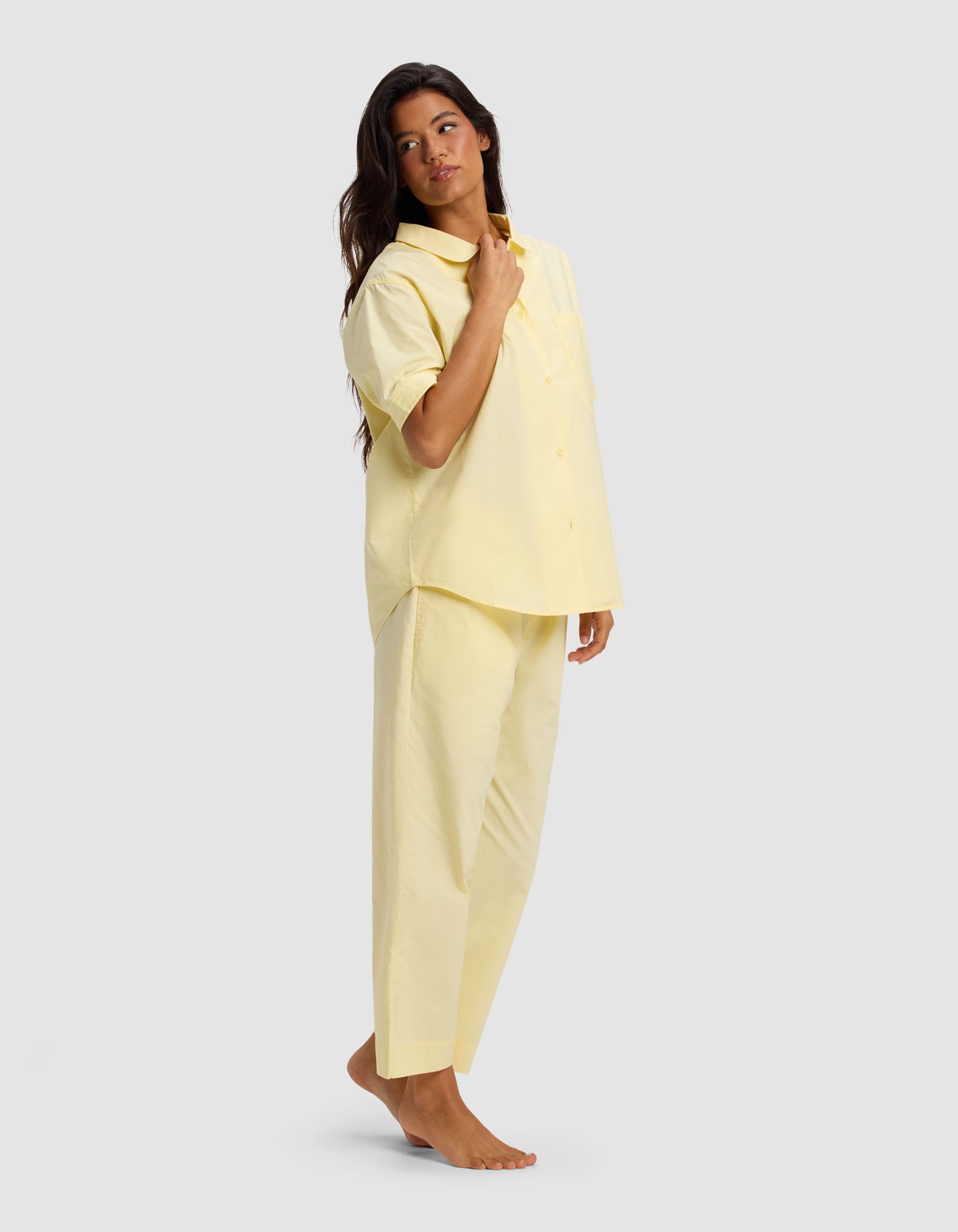 A woman stands barefoot against a plain background, wearing the Cozy Earth Women's Sutton Short Sleeve & Cropped Pant Pajama Set in light yellow. She looks slightly to the side and touches her collar with one hand.|Color:Meringue
