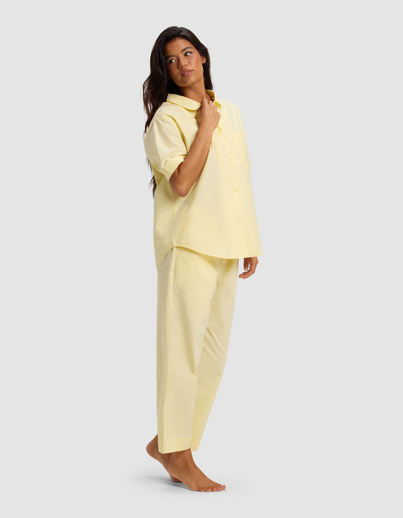 A woman stands barefoot against a plain background, wearing the Cozy Earth Women's Sutton Short Sleeve & Cropped Pant Pajama Set in light yellow. She looks slightly to the side and touches her collar with one hand.
