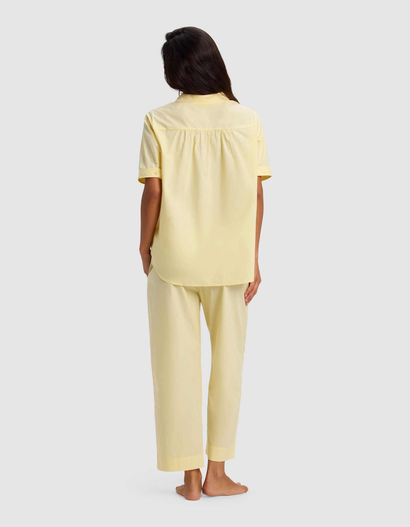 A person stands barefoot, facing away, wearing the Cozy Earth Women's Sutton Short Sleeve & Cropped Pant Pajama Set in light yellow. The background is plain and light gray.