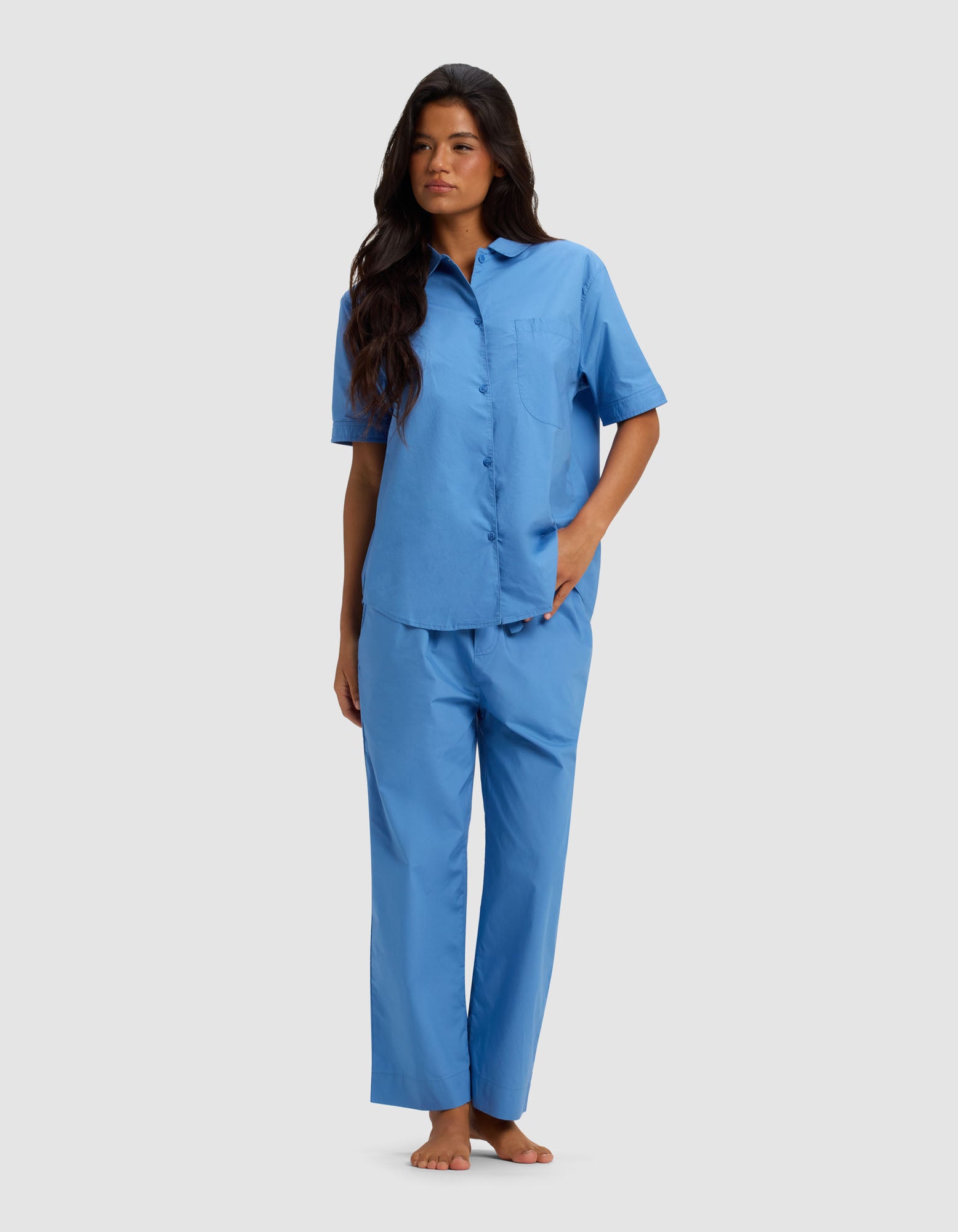A woman with long dark hair stands barefoot against a plain background, wearing the Cozy Earth Women's Sutton Short Sleeve & Cropped Pant Pajama Set in light blue, with one hand in her pocket and gazing slightly to the side.|Color:Sailor