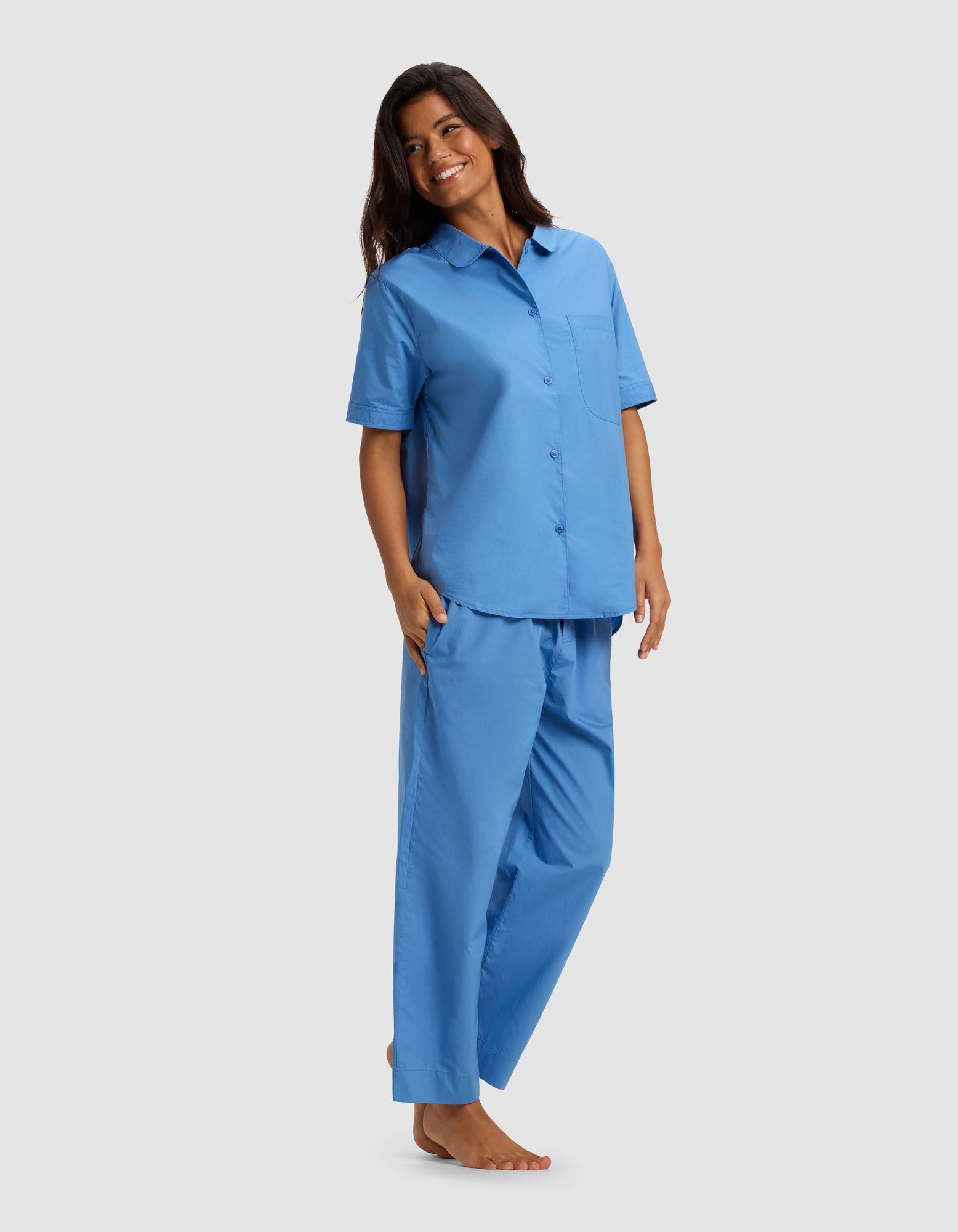 A woman with long dark hair smiles in the Cozy Earth Women's Sutton Short Sleeve & Cropped Pant Pajama Set—loose-fitting, light blue pajamas featuring short sleeves and cropped pants—standing barefoot against a plain, light gray background.