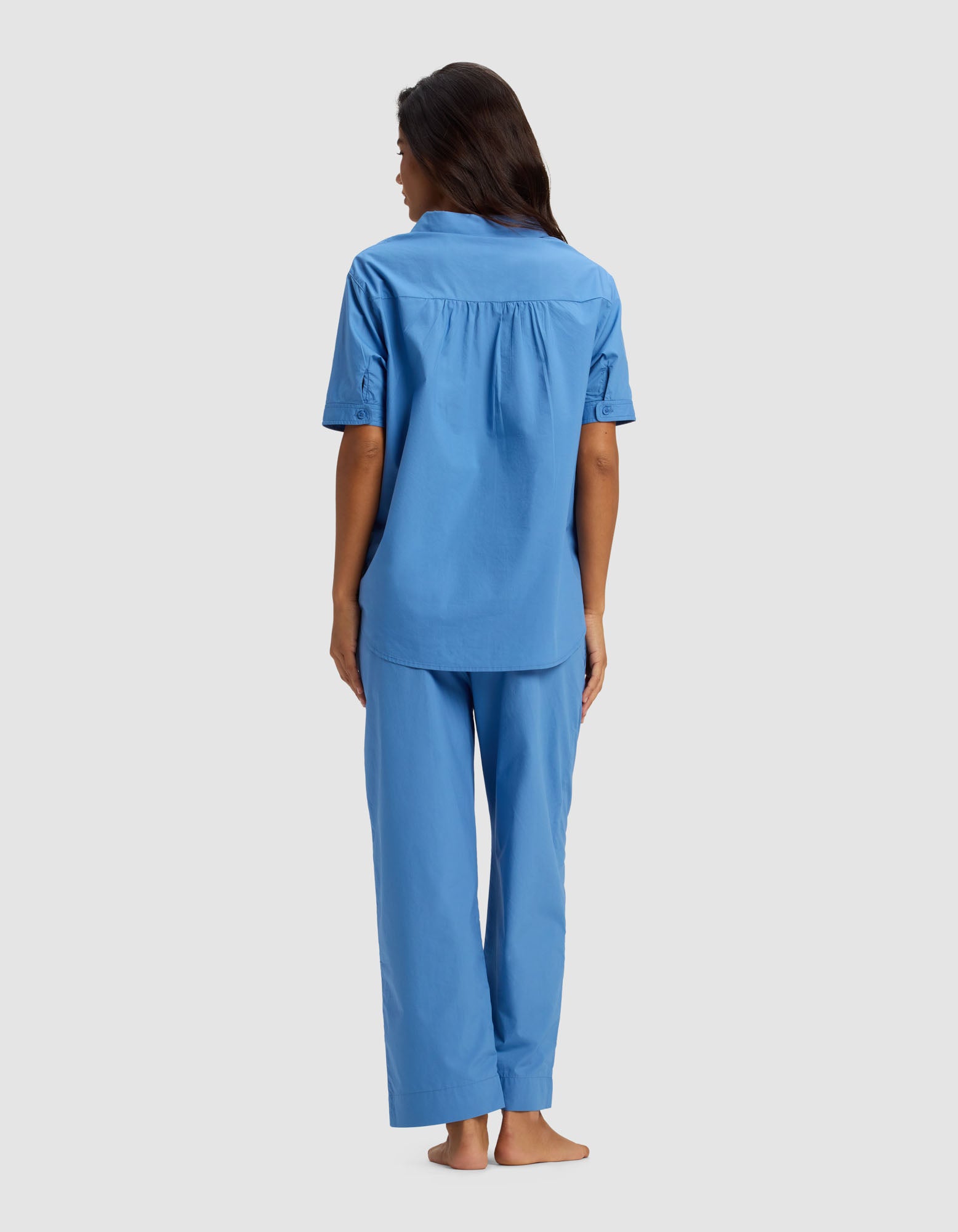 A woman stands barefoot facing away, wearing the Cozy Earth Women's Sutton Short Sleeve & Cropped Pant Pajama Set in light blue, against a plain light gray background.