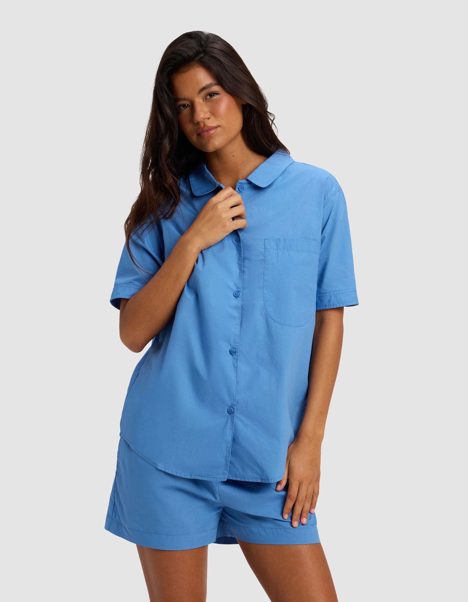A woman with long brown hair wears the Cozy Earth Women's Sutton Short Sleeve Pajama Top in light blue, standing against a plain light gray background. She touches her collar with one hand and rests the other by her side.|Color:Sailor