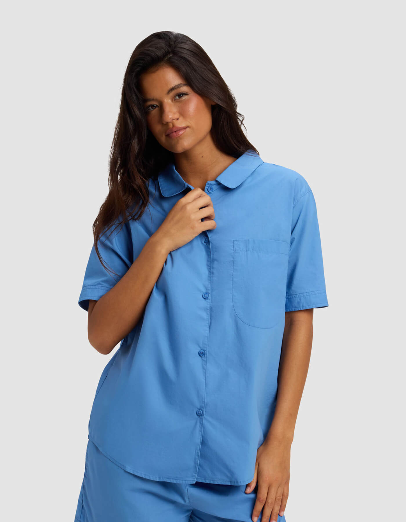 A woman with long dark hair wears the Cozy Earth Women's Sutton Short Sleeve & Cropped Pant Pajama Set, standing against a plain light gray background. She looks relaxed and holds her shirt collar with one hand.
