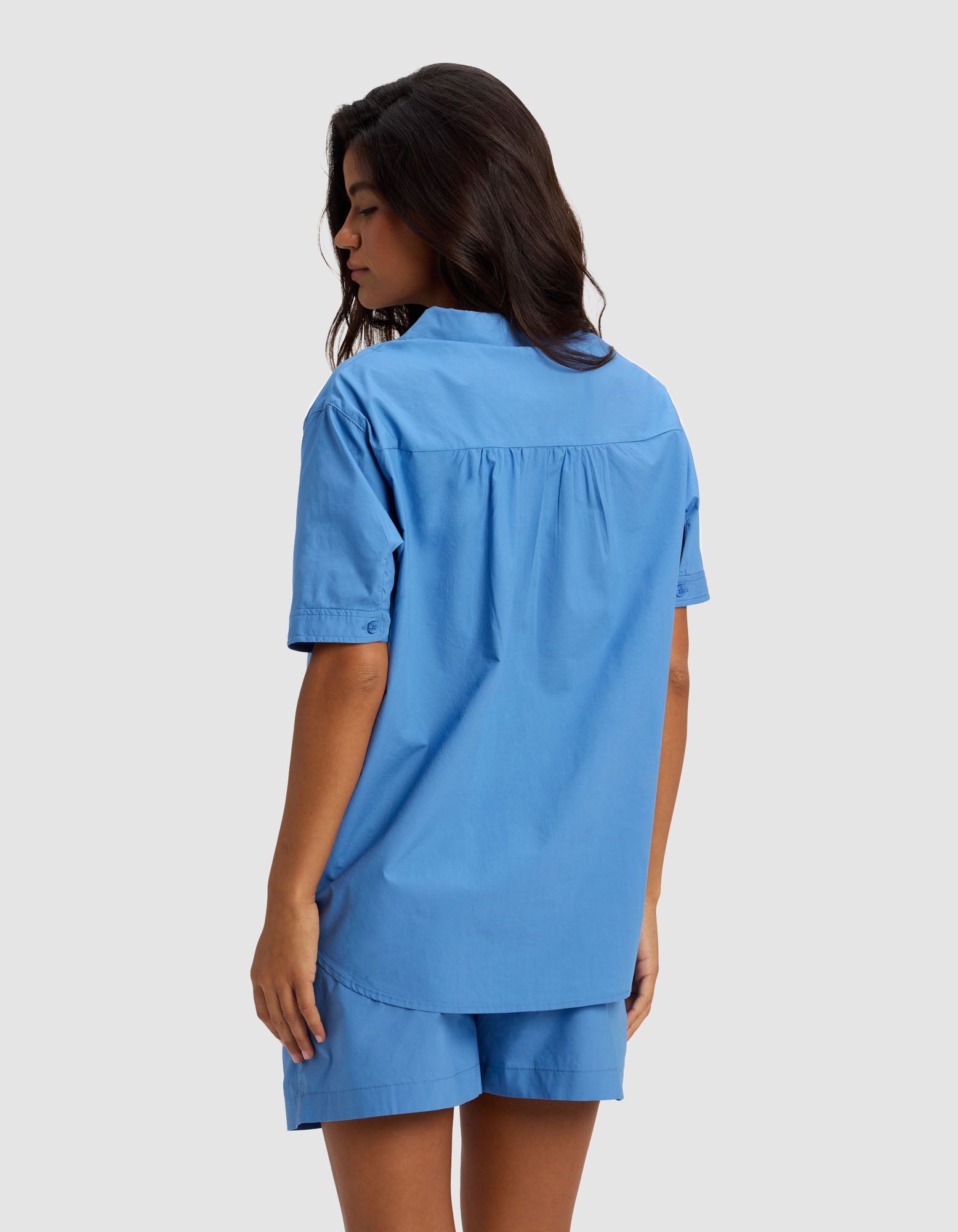 A woman with long dark hair stands sideways, facing away, wearing the Cozy Earth Women's Sutton Short Sleeve Pajama Top in blue and matching shorts against a plain light gray background.